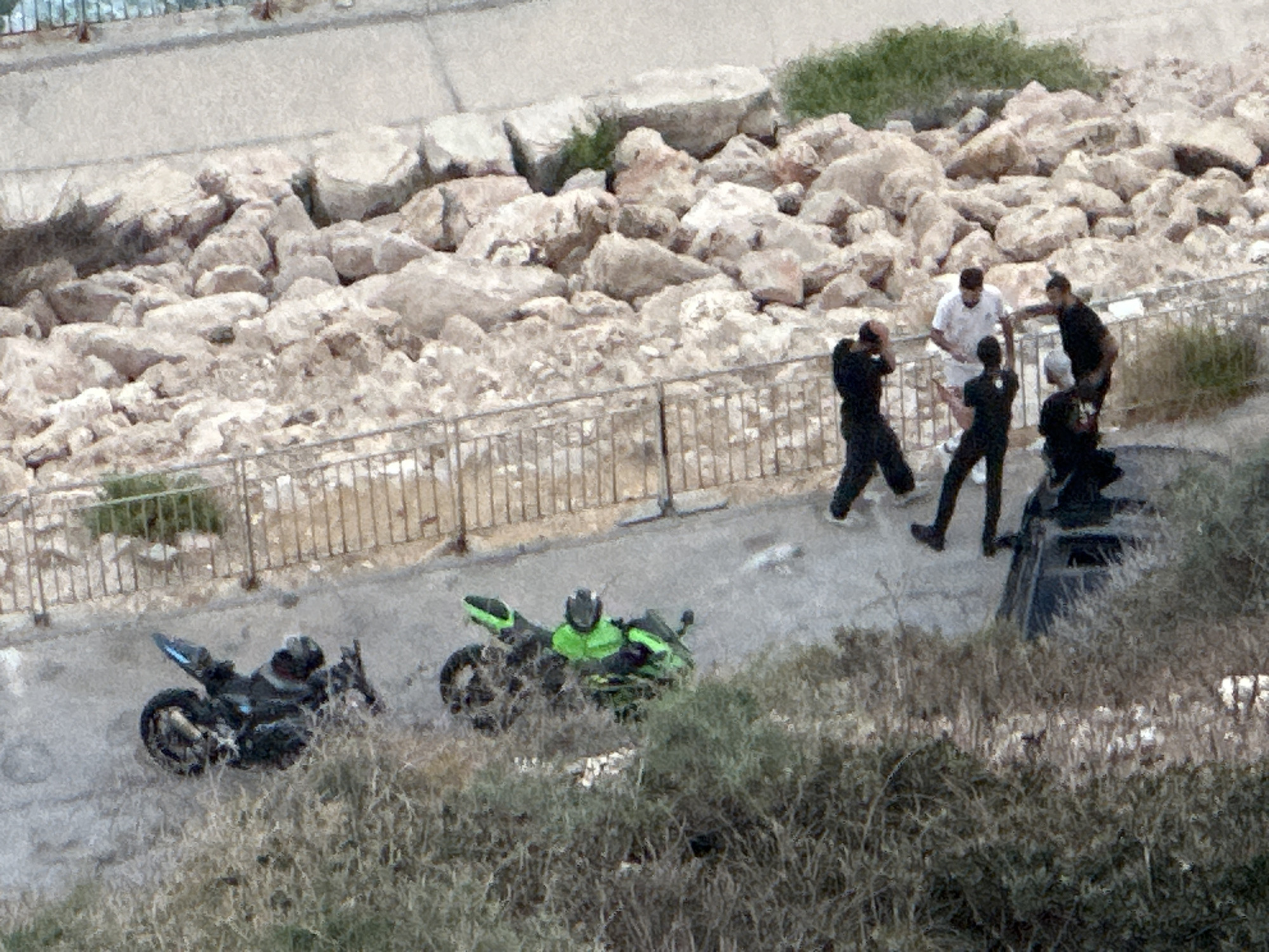 In Mate Asher Regional Council, Israel, an outdoor scene captures a group of individuals near a rocky area and a paved path. Two motorcycles are parked on the path, one black and one green. Several men are gathered, some appearing to be interacting with each other. One man in a white shirt stands with his hand extended. Other men are dressed in dark clothing, with one in a black outfit appearing to be on the ground. The environment includes a metal fence, a grassy area, and a large pile of light-colored rocks.
