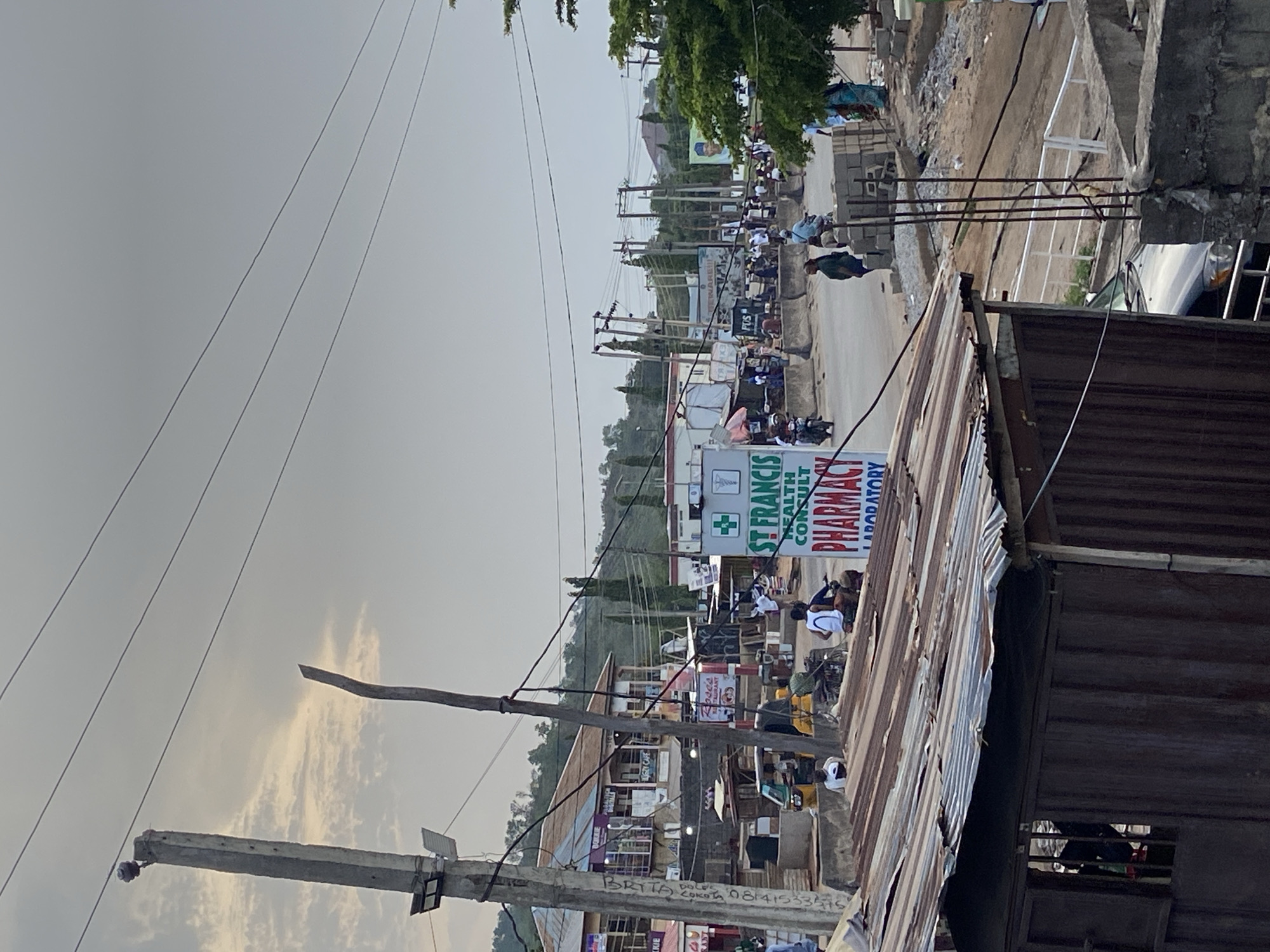 The image shows a street scene in Lokoja, Nigeria. The angle is slightly oblique. In the background, a building is visible with a sign for "St Francis Health Consult Pharmacy Laboratory." People, some wearing white shirts, are gathered on the street. Power lines are strung across the sky. In the foreground, a corrugated metal roof obscures part of the view, and the frame of a small wooden structure or building is on the right. A concrete utility pole is visible on the left, with exposed wiring. The sky is overcast.