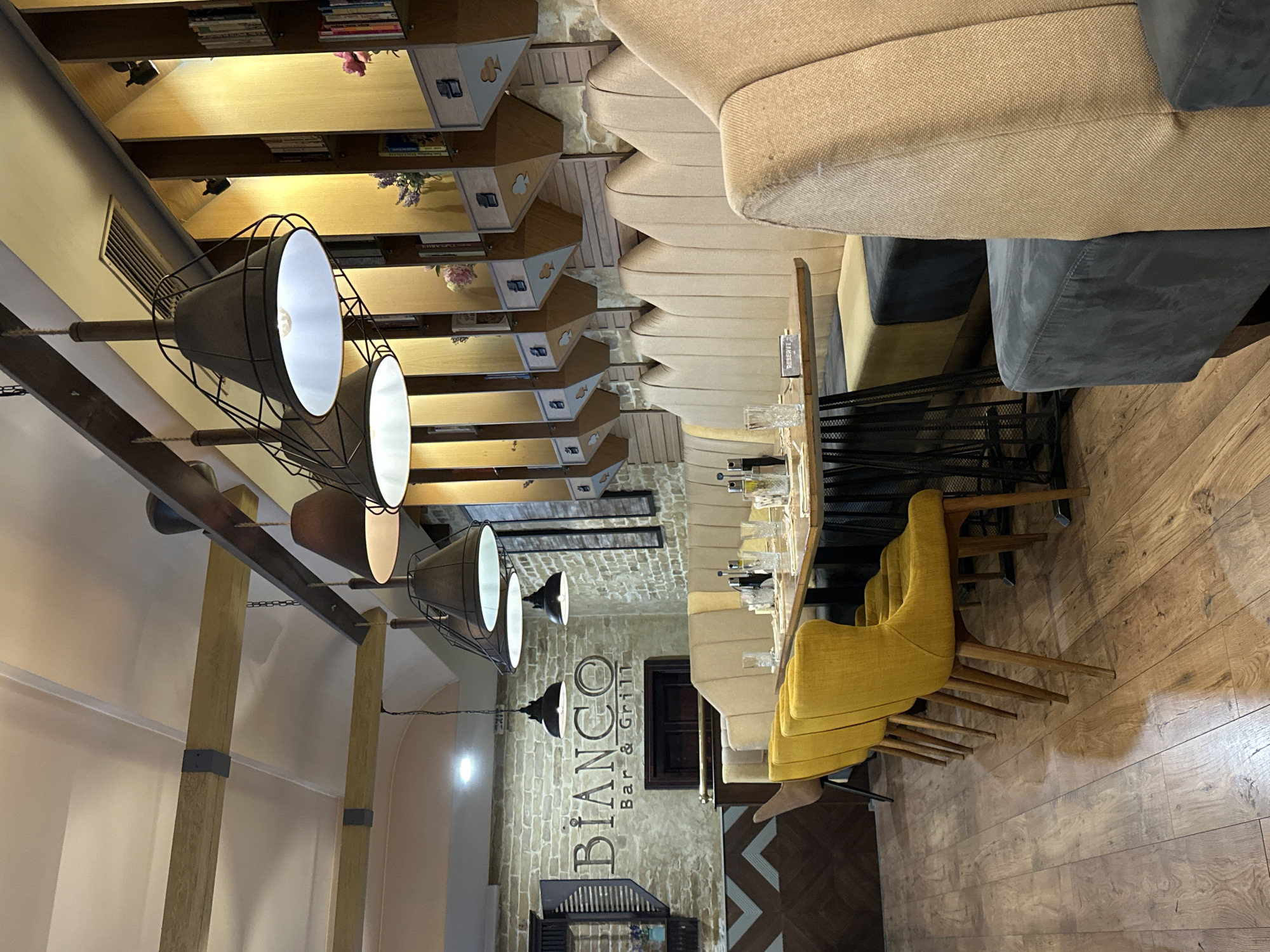 The interior of a restaurant, named "Bianco Bar & Grill", is visible. The restaurant is located in Veliko Tarnovo, Bulgaria. The scene is dominated by a long table with multiple yellow chairs. A large, multi-lamp lighting fixture hangs overhead. On the opposite wall, rows of shelves are visible. The seating area appears to have a beige couch. The floor is a wooden material.
