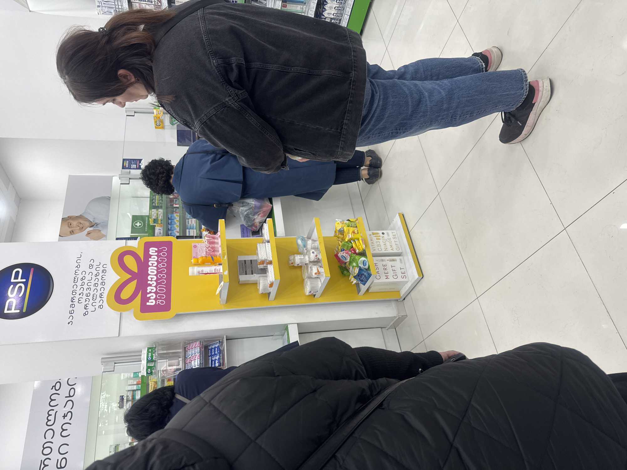 The image captures an indoor scene inside a pharmacy or a health and beauty store, identifiable by the "PSP" logo and a green pharmacy cross symbol. The setting is bright with white walls and a light-colored tiled floor, consistent with a modern retail environment. The presence of Georgian text on signs confirms the location as Tbilisi, Georgia.
In the foreground, a woman with dark hair tied back, wearing a dark denim jacket, blue jeans, and black sneakers with pink accents, is looking downwards, seemingly examining products on a lower shelf. Behind her, another individual in a dark blue jacket is similarly focused on items. A third person in a black quilted jacket is partially visible in the lower right.
The store features various display shelves and a prominent yellow stand filled with small packaged goods. This stand displays Georgian text meaning "Best offer," alongside English phrases like "CASH MERE GIFT SET" and "VITAMIN C.B3 NIACIN AMIDE GIFT SET." Product packaging, such as "Orbit-it" gum, is also visible. Large posters on the walls promote the "PSP" brand, with Georgian text that translates to "In the environment of health, care, and beauty," and one poster includes an image of a smiling man. The activity taking place appears to be customers browsing or shopping.