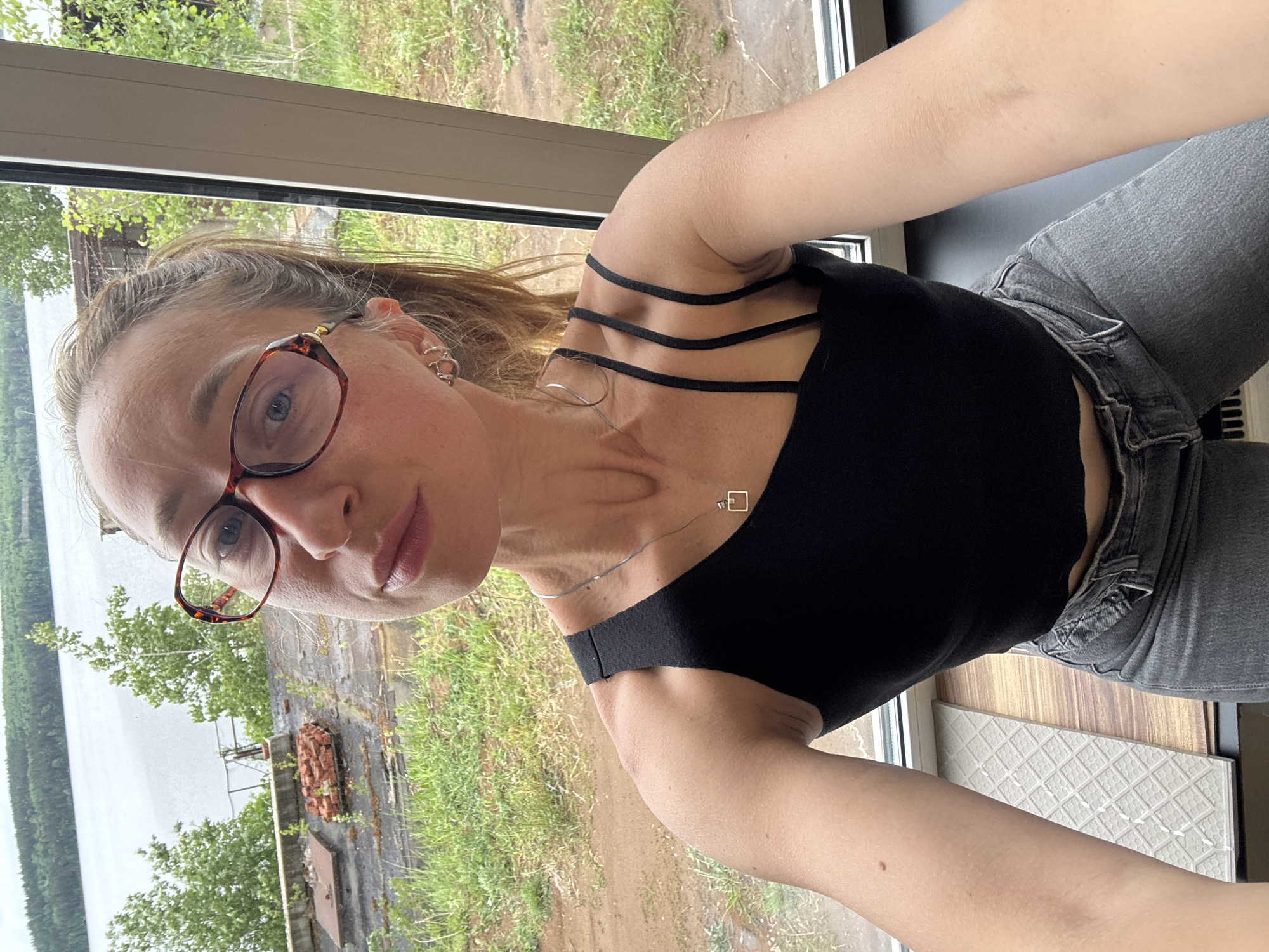 A woman wearing a black tank top, grey jeans, and glasses poses indoors, likely in a residential building, as a view from the window shows an exterior landscape of trees, grass, a body of water, and what appears to be an abandoned structure with exposed brick. The location is in Chaikovsky, Russia. The woman wears jewelry including a necklace and earrings.