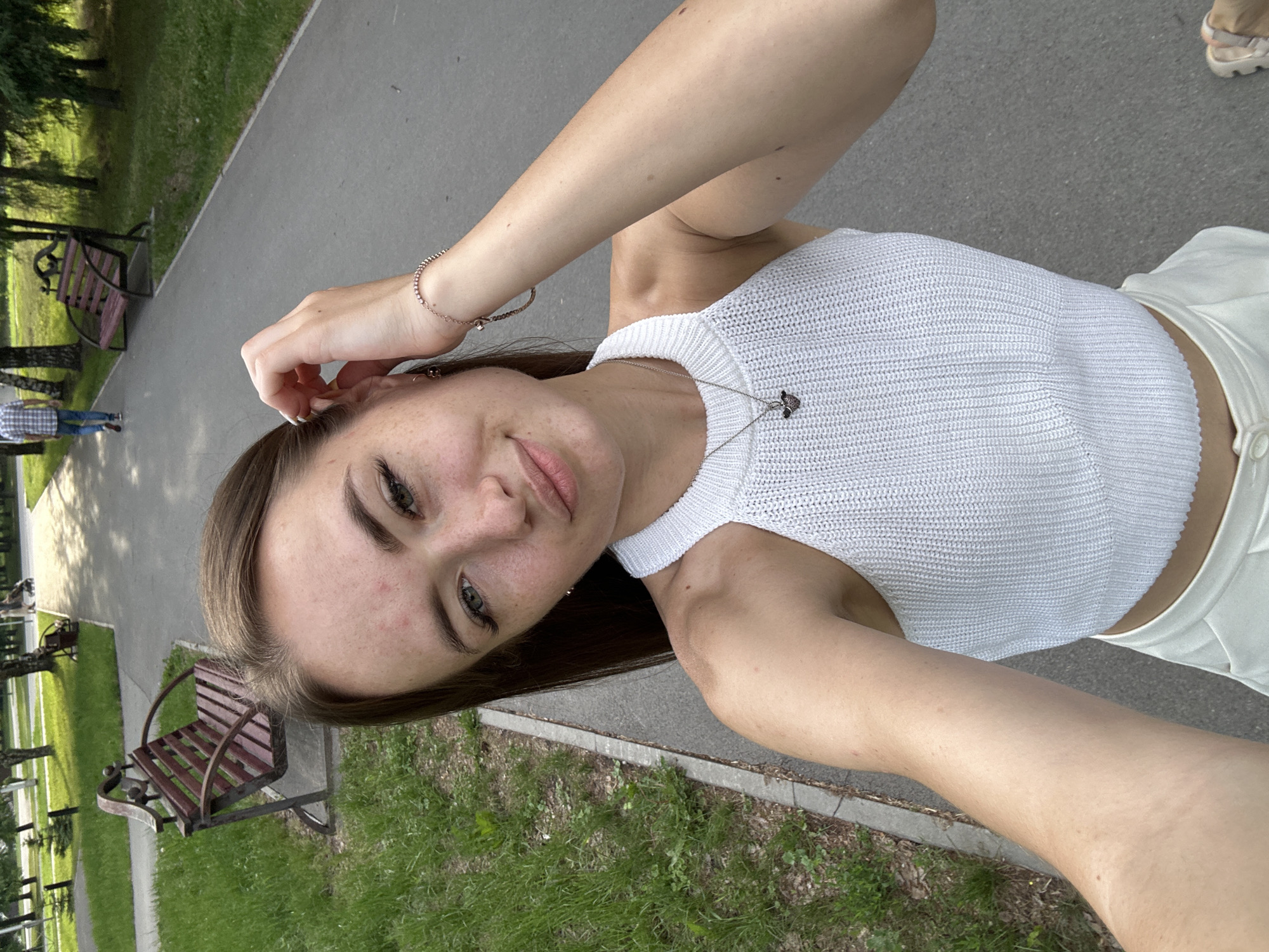 A young woman is taking a selfie outdoors in a park. She has long brown hair, freckles, and is wearing a white tank top and white pants. She is touching her hair with her right hand. In the background, there are trees, a paved path, and park benches. One person is walking away from the camera in the distance. The lighting suggests it is daytime.