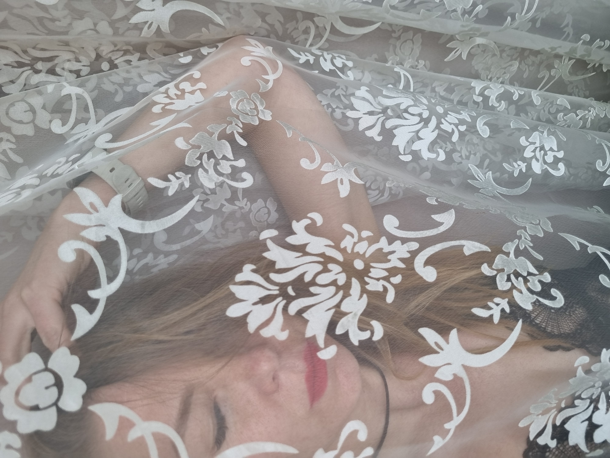 A close-up, high-angle shot captures a person, likely a woman, lying beneath a translucent white fabric adorned with an intricate, damask-style floral pattern. The sheer material partially obscures the individual, creating a soft, ethereal effect.
The person's face is visible through the fabric, with eyes closed, suggesting a state of rest or contemplation. Bright red lipstick is applied to her lips. Her light-colored hair, possibly blonde or light brown, is spread out around her head. Her arms are bent, with one arm extended above her head. A light-colored wristband or watch is visible on her left wrist. On her right side, a glimpse of a dark, possibly lacy garment, can be seen beneath the patterned fabric.
The image is tightly framed, emphasizing the delicate interplay between the patterned textile and the figure beneath. The lighting appears soft and even, contributing to the gentle aesthetic. There is no visible text in the image. While the location context provided is Chaikovsky, Russia, the visual content of the image does not offer specific cues about this location.