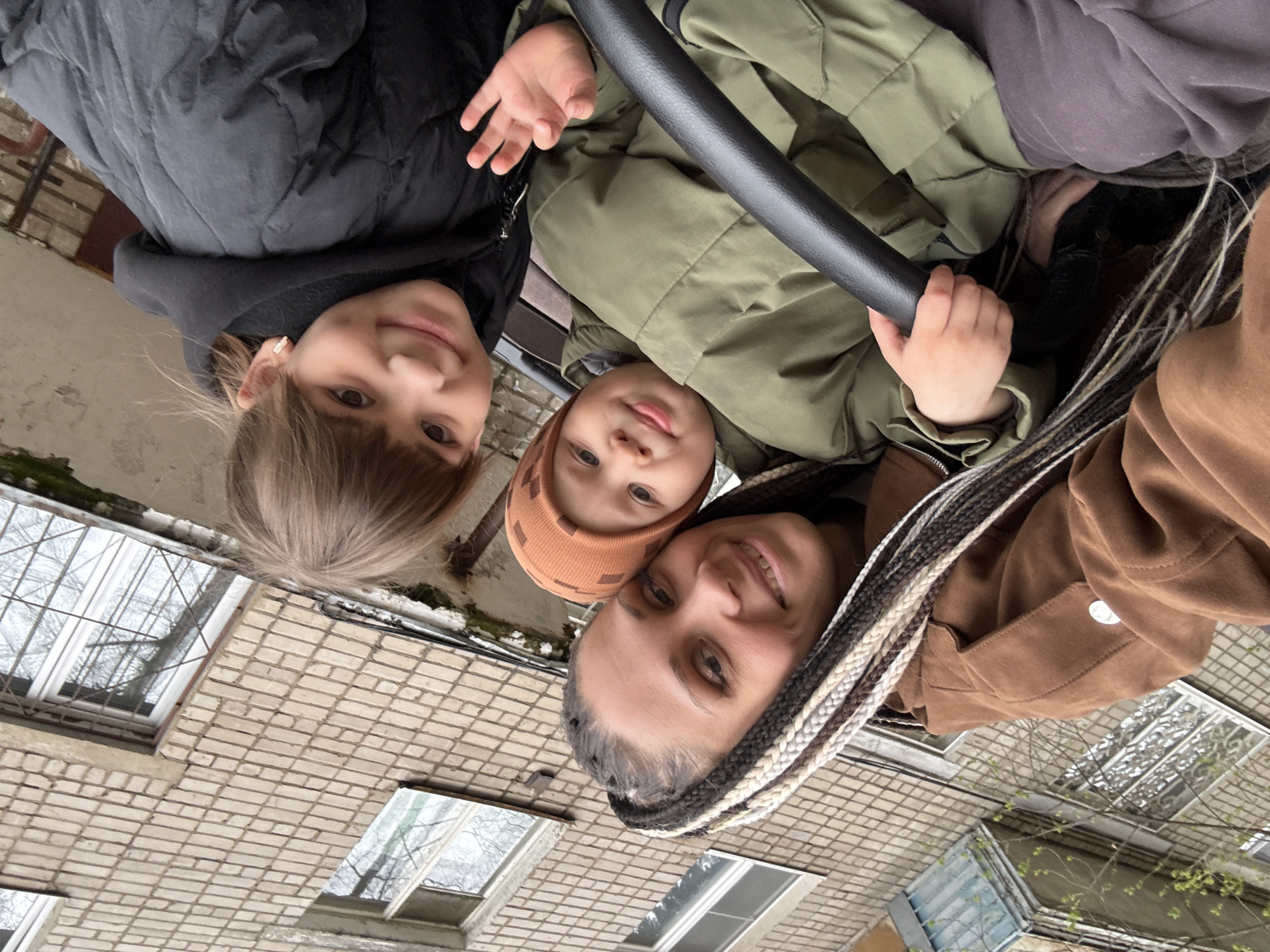 Three people, including two children, are posing for a selfie or photograph together. The orientation of the picture suggests the camera is angled downwards, looking up towards the subjects. The backdrop features a building constructed of light-colored bricks. One child, on the left, wears a black jacket, while the other child is wearing an olive green jacket and an orange beanie. The adult on the right has braided hair and wears a brown jacket. The photo was taken in an urban setting, likely in Rusanovo, Russia, as inferred from the surrounding architecture.