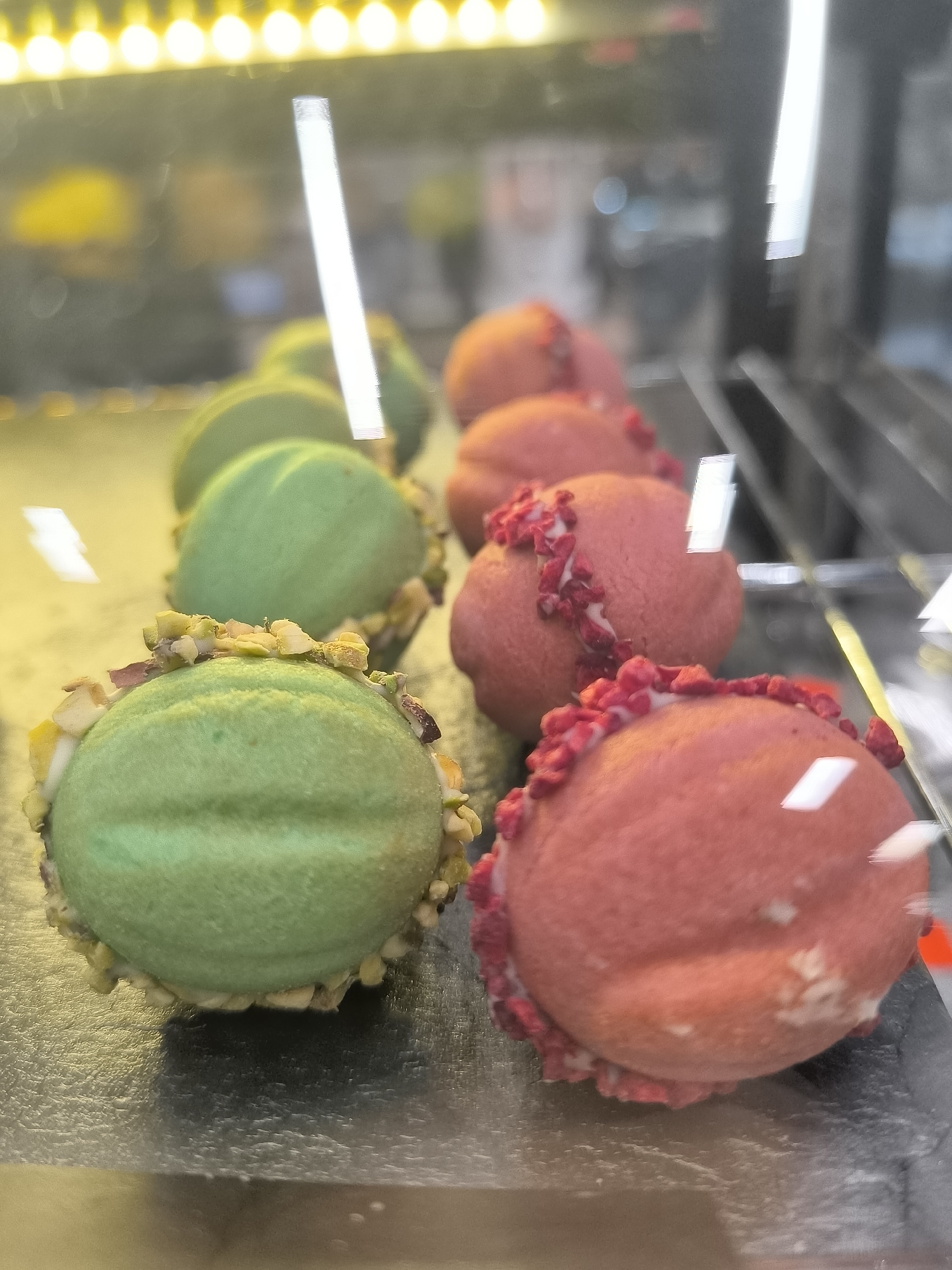 This image shows a close-up, slightly angled view of brightly colored pastries, likely macarons, displayed in a refrigerated case. Two green pastries coated with chopped nuts are in the foreground, with several pink pastries adorned with red, dried fruit pieces visible behind them. The pastries are arranged in rows on a reflective surface, possibly a glass shelf, within the case. Above the pastries, bright yellow and white lights create a warm, ambient glow, reflected in the glass. The background is blurred, suggesting a retail environment, possibly a bakery or cafe in Dnipro, Ukraine. The overall impression is one of vibrant colors and tempting desserts.