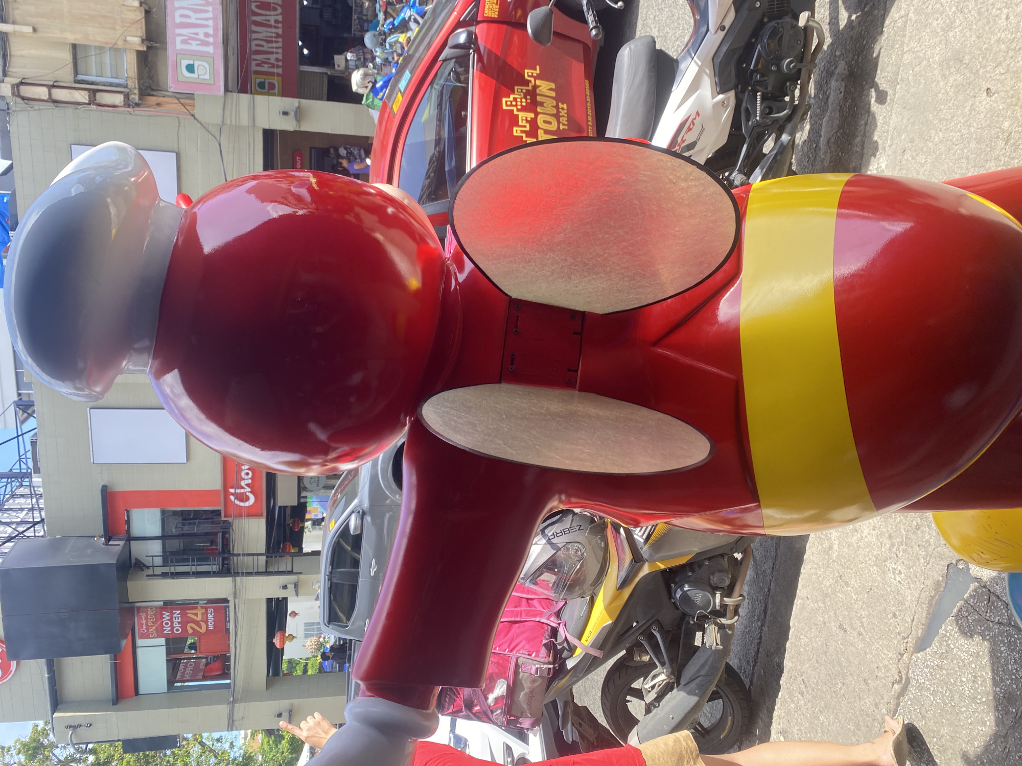 A large, bright red and yellow anthropomorphic bee mascot, likely from Jollibee, dominates the foreground of a sunlit urban street scene. The mascot wears a white chef's hat and has large, light-colored circular ears, with a distinctive yellow stripe around its red body. Partially visible in the bottom left are a person's arm, pointing towards the background, and a leg.
Behind the mascot, a red taxi, prominently labeled "TOWN TAXI" and bearing the registration "CITY R-XI 2019-XI-00500," is parked on the street. Adjacent to the taxi are several motorcycles, one yellow and black with a visible "foodpanda" delivery bag, and another white and black. A helmet with "ZEBRA" written on it rests on one of the bikes. A gray car is also partially visible further back.
The background features multi-story buildings housing various businesses. A "Chowking SAN PEDRO" fast-food restaurant is clearly visible, advertising "NOW OPEN 24 HOURS." Further down the street, a "FARMACIA" sign can be seen on another building. The architecture suggests a commercial district, with concrete structures and multiple storefronts.
The time of day is clearly daytime, characterized by bright sunlight casting distinct shadows, indicating clear weather. The combination of the Jollibee mascot, Chowking restaurant, and the Davao City taxi markings strongly places the scene in the Philippines, specifically Davao City, as indicated by the context. The street appears active with vehicles and a few distant pedestrians.