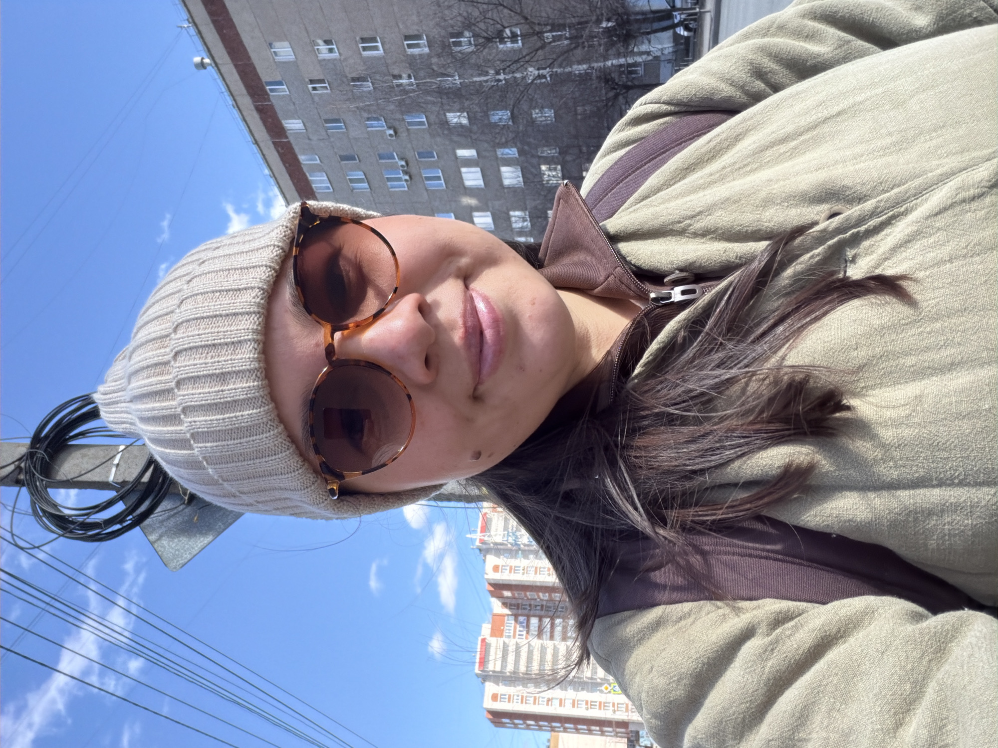 A woman is taking a selfie. She is wearing a beige beanie and sunglasses. The building in the background is likely located in Izhevsk, Russia. There are overhead wires.