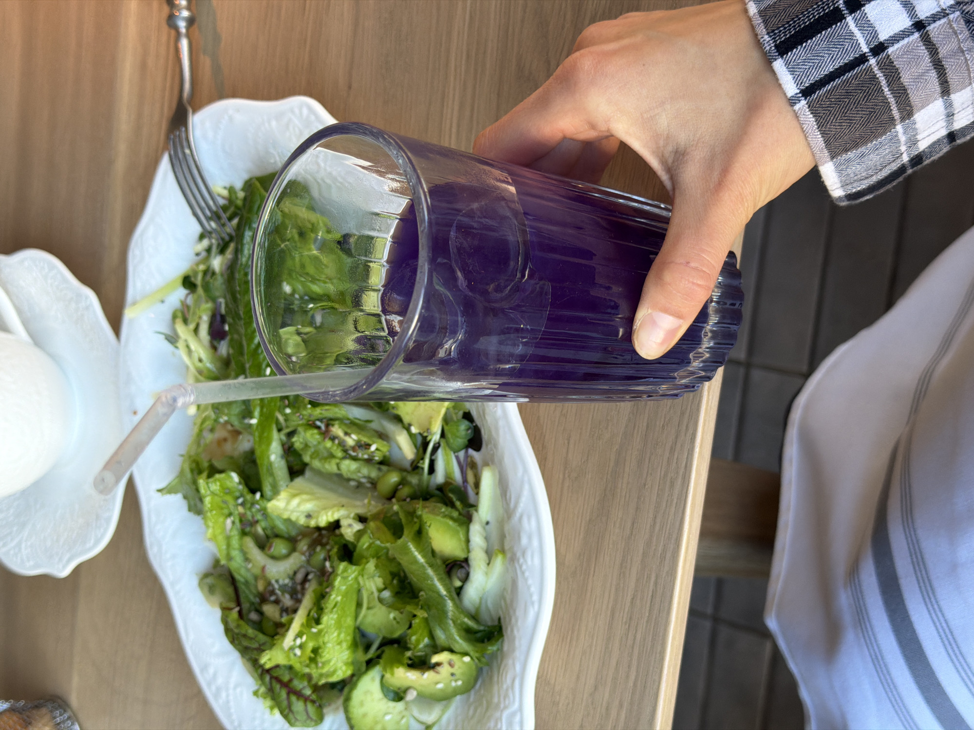 A close-up, elevated shot shows a person pouring a purple liquid from a tall glass into a white, ornate bowl of salad. The salad contains green leaves, avocado, and other ingredients. A fork is positioned in the bowl, and a straw extends from near the white saucer on the side into the salad. The person pouring is wearing a black and white plaid shirt, and a white striped towel is visible on the right side of the image. The setting appears to be on a wooden table. No information can be discerned that indicates the location is Krasnoyarsk, Russia.