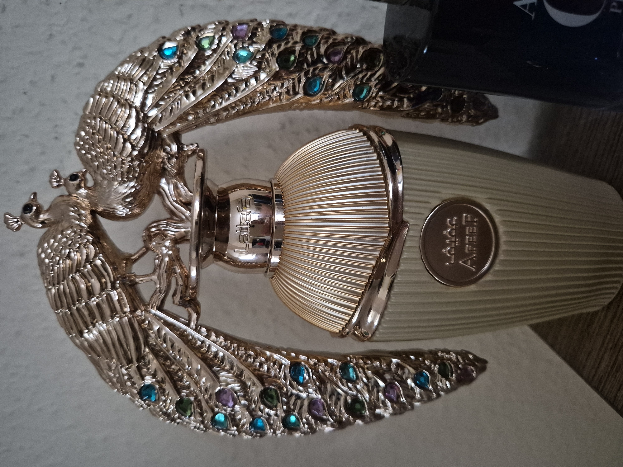 The image shows a close-up of a perfume bottle with an ornate, gold-colored peacock design. Two stylized peacocks, adorned with colorful gemstones in blue, green, and purple, arch over the bottle's cap. The bottle itself is a pale beige with vertical ribbed detailing and a gold band near the top. A circular emblem with Arabic script and the word "AFEEF" is visible on the front of the bottle. The background is a plain, textured wall. The scene is lit from above and to the left, casting shadows. To the right of the perfume bottle, partially visible, is another dark-colored bottle with white lettering, including the letters "A" and "O". The perfume bottle is placed on a wooden surface.