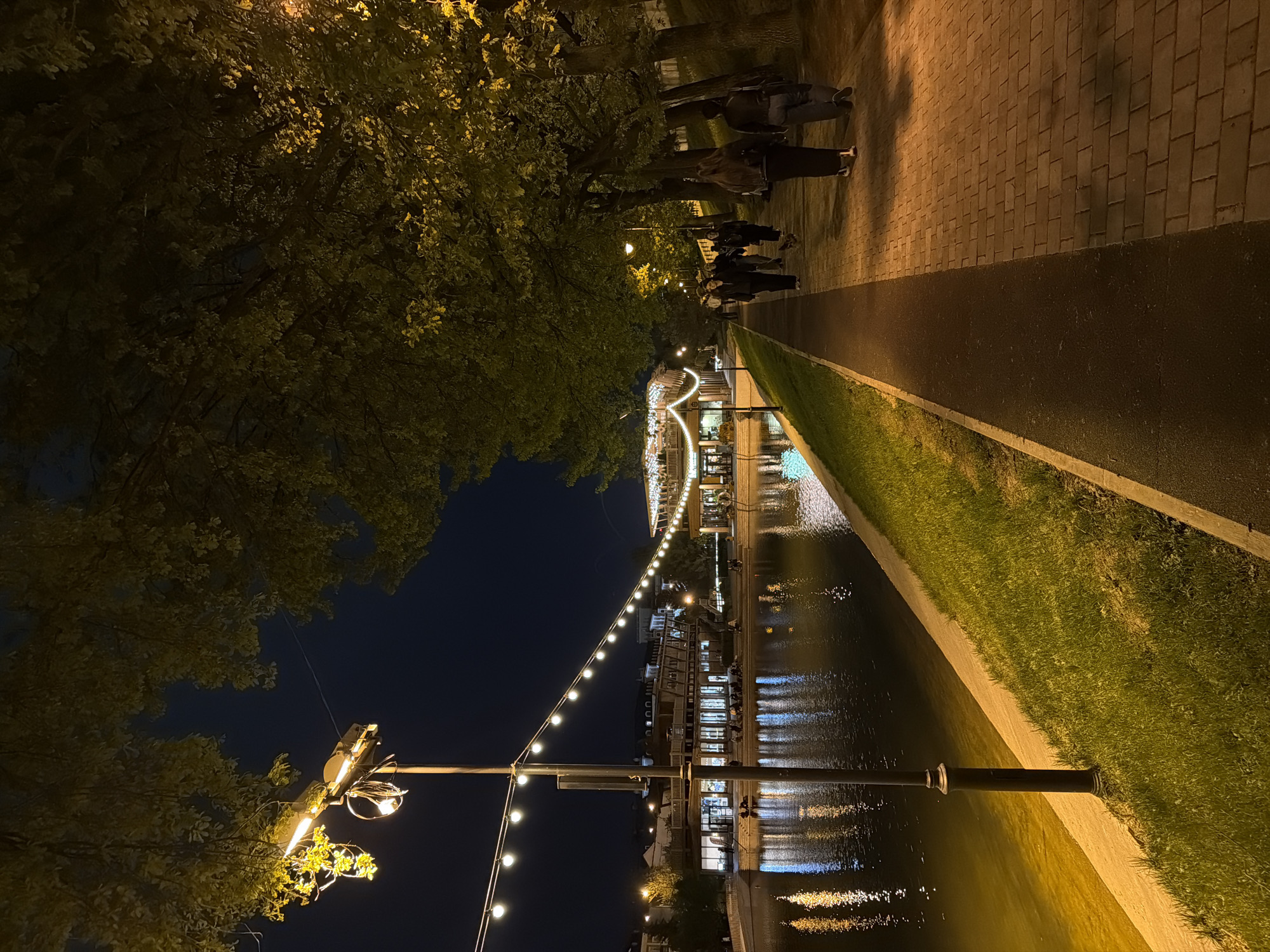 The image depicts a night scene along a canal in Kazan, Russia. A brick walkway and paved path run parallel to the canal, lined with grass. Several people are walking along the path. Strings of lights illuminate the scene and reflect in the water. Trees border the walkway. A building with illuminated arches and reflections is visible in the background. A streetlight stands in the foreground.