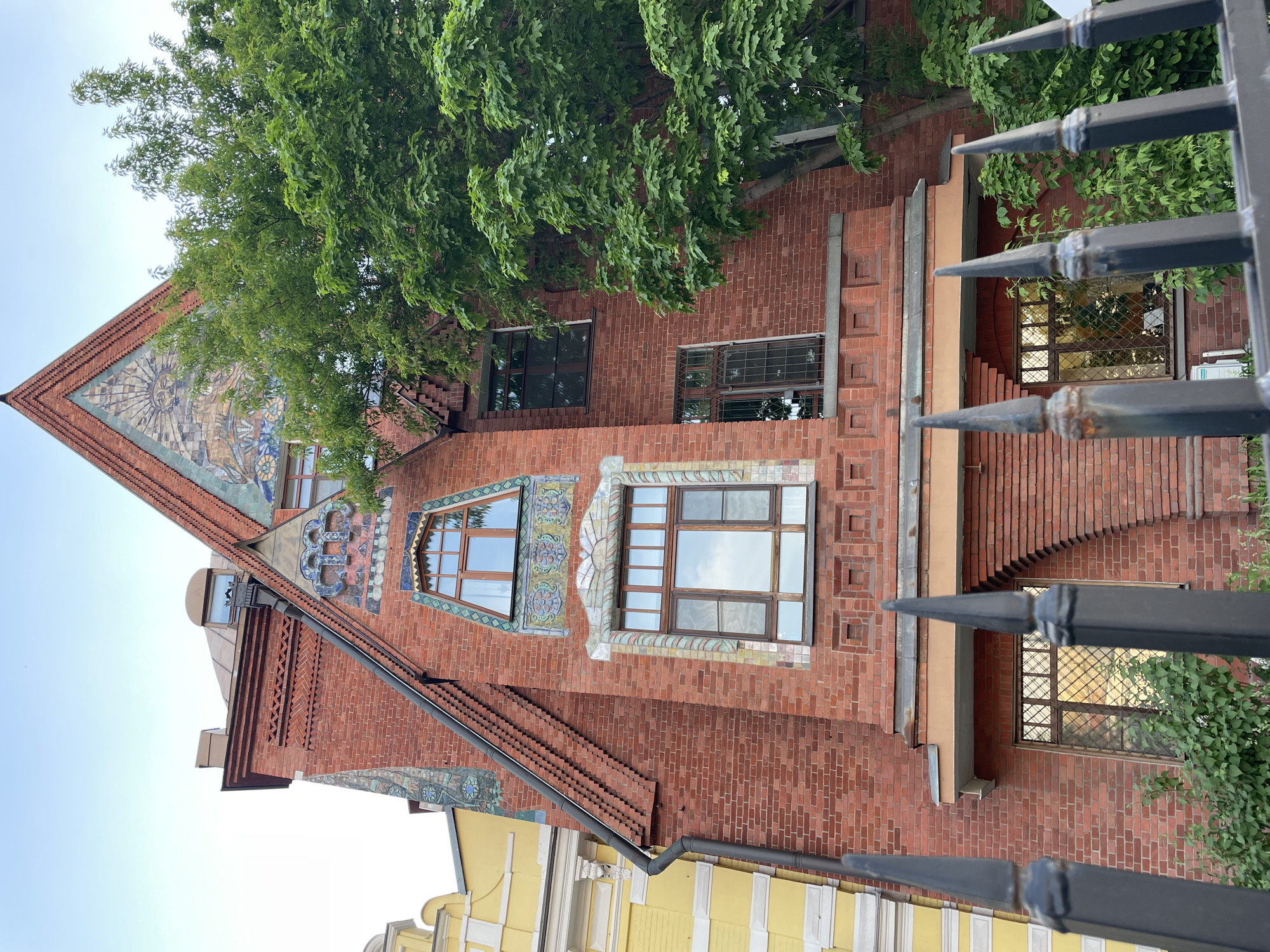 The image shows a building with a unique architectural style in an urban environment. The building is primarily red brick, with decorative mosaic tilework around the windows and on the upper section of the triangular roof. The windows are framed in wood. A large green tree partially obscures the upper portion of the building. A metal fence with pointed finials is visible in the foreground, slightly out of focus. The sky is a pale blue. A yellow building is seen below. The location is Unknown, Unknown.