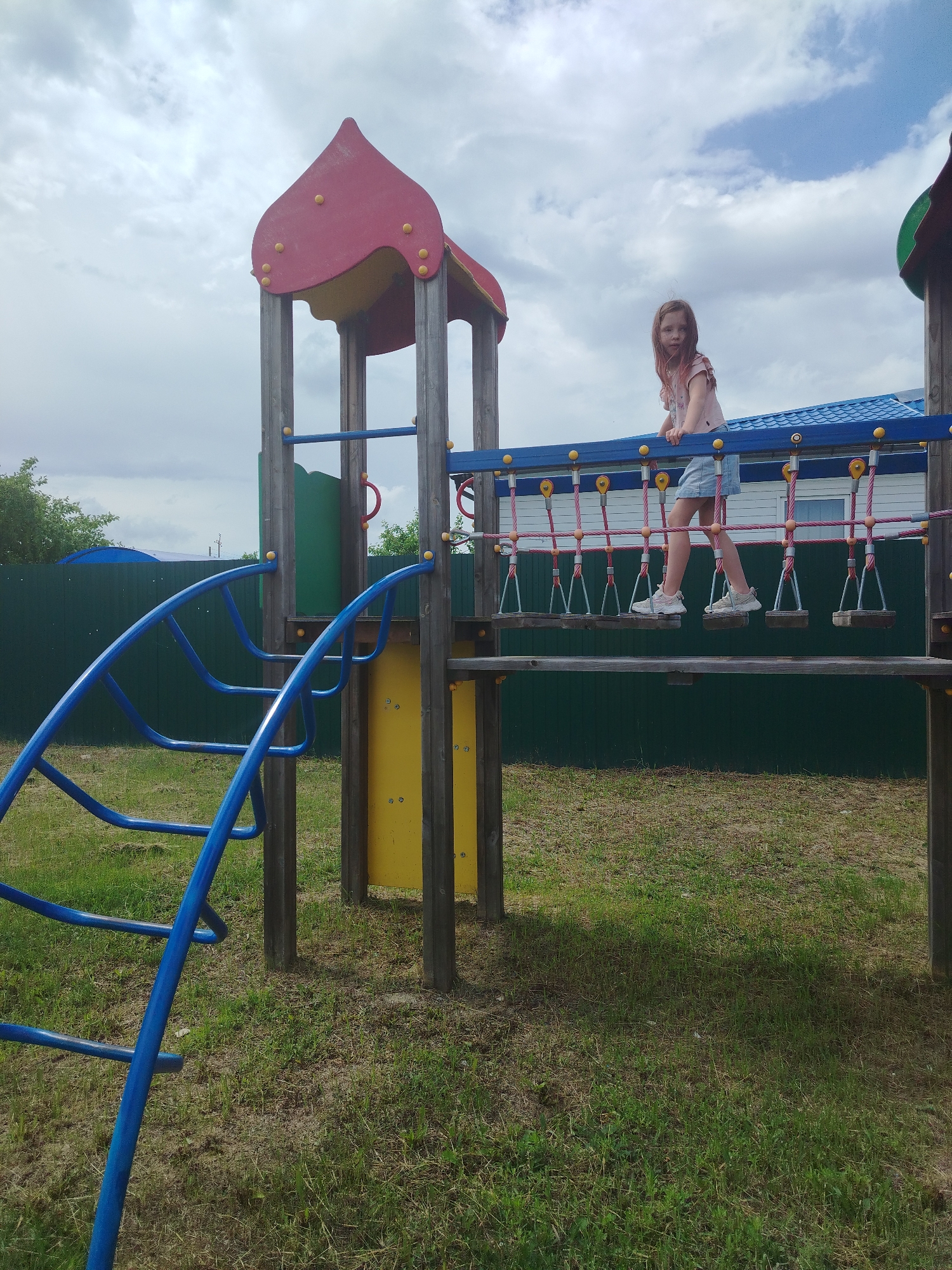 In Klyuchi, Russia, on a cloudy day, a young girl with long auburn hair stands on a playground bridge. She is wearing a light pink t-shirt and a denim skirt. The bridge is made of rope and wooden planks, suspended between two wooden play structures. One play structure has a red, curved roof with yellow decorative accents. A blue metal slide is visible on the left side of the image, curving upwards. The playground is situated on a patch of dry, sparse grass with some green patches. In the background, a dark green fence runs along the property line, and a building with a blue roof is partially visible. The sky is filled with grey and white clouds.