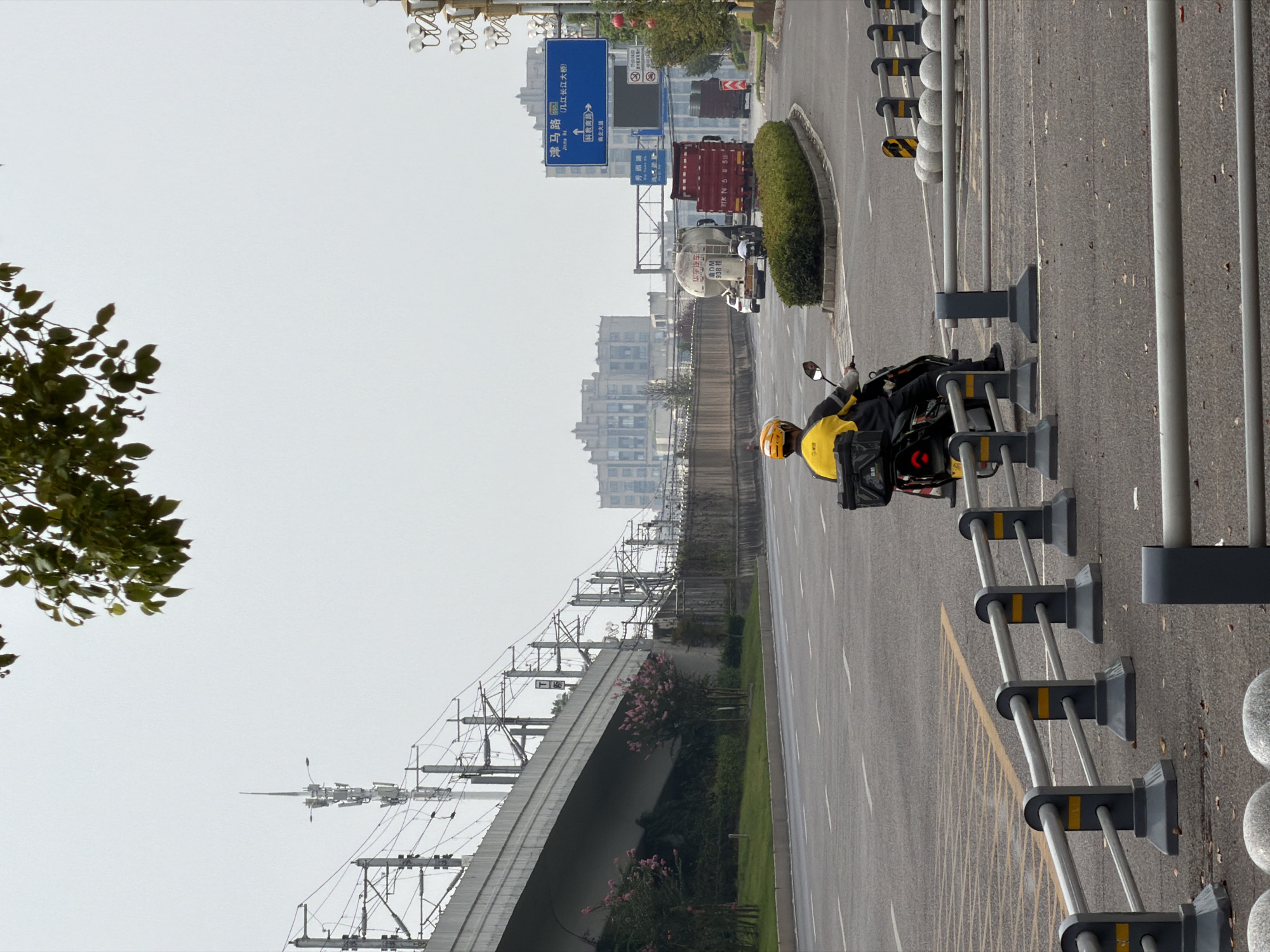 The image depicts an outdoor urban scene in Jiangjin, China, characterized by a slightly overcast sky and a muted color palette. In the foreground, a person wearing a yellow jacket and helmet rides a scooter, equipped with a black delivery box on the rear. The scooter is on a paved road with white lane markings. Various traffic barriers made of gray metal and concrete line the road.
In the midground, several large vehicles are visible, including a white tanker truck and a red truck. Traffic signs are present, with one blue sign displaying Chinese text and directional arrows. The road curves and extends into the distance, bordered by a raised concrete barrier and a line of greenery.
The background features a large, multi-story building with a distinctive architectural style, situated behind a series of elevated structures and electrical pylons. A complex network of overhead power lines and communication towers is prominently displayed, suggesting the presence of significant infrastructure. Trees with pink blossoms are planted along the roadside, adding a touch of color to the scene. The overall impression is of a busy urban environment with a focus on transportation and infrastructure.