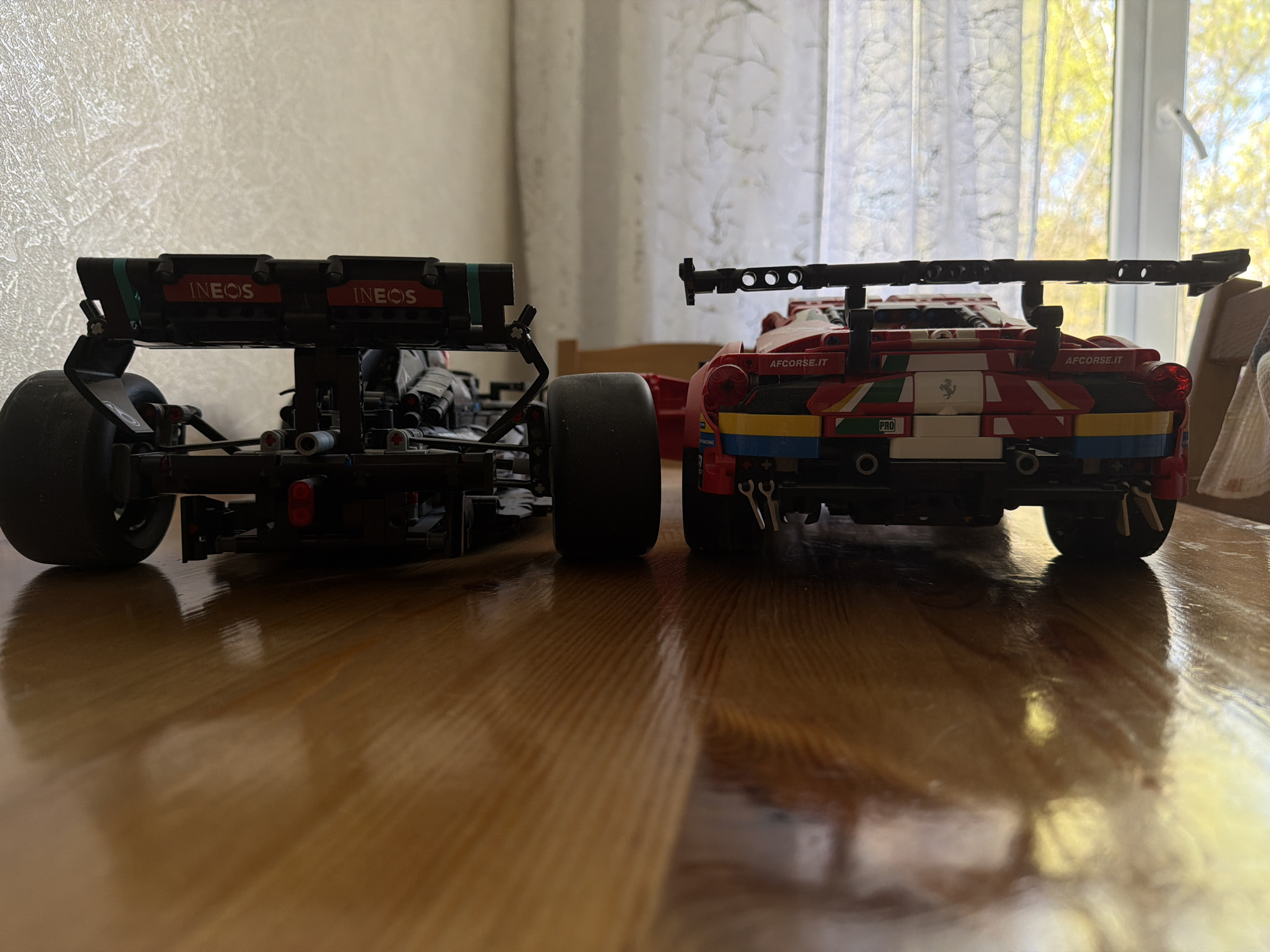 The image shows two LEGO Technic model cars from the rear, standing on a varnished wooden surface, likely a table. The car on the left is black and has "INEOS" printed on its rear wing. The car on the right is primarily red and has "AFCorsa.it" printed on it. A window with thin white curtains is visible in the background. The image does not offer evidence to corroborate the location in Pavlovo, Russia.