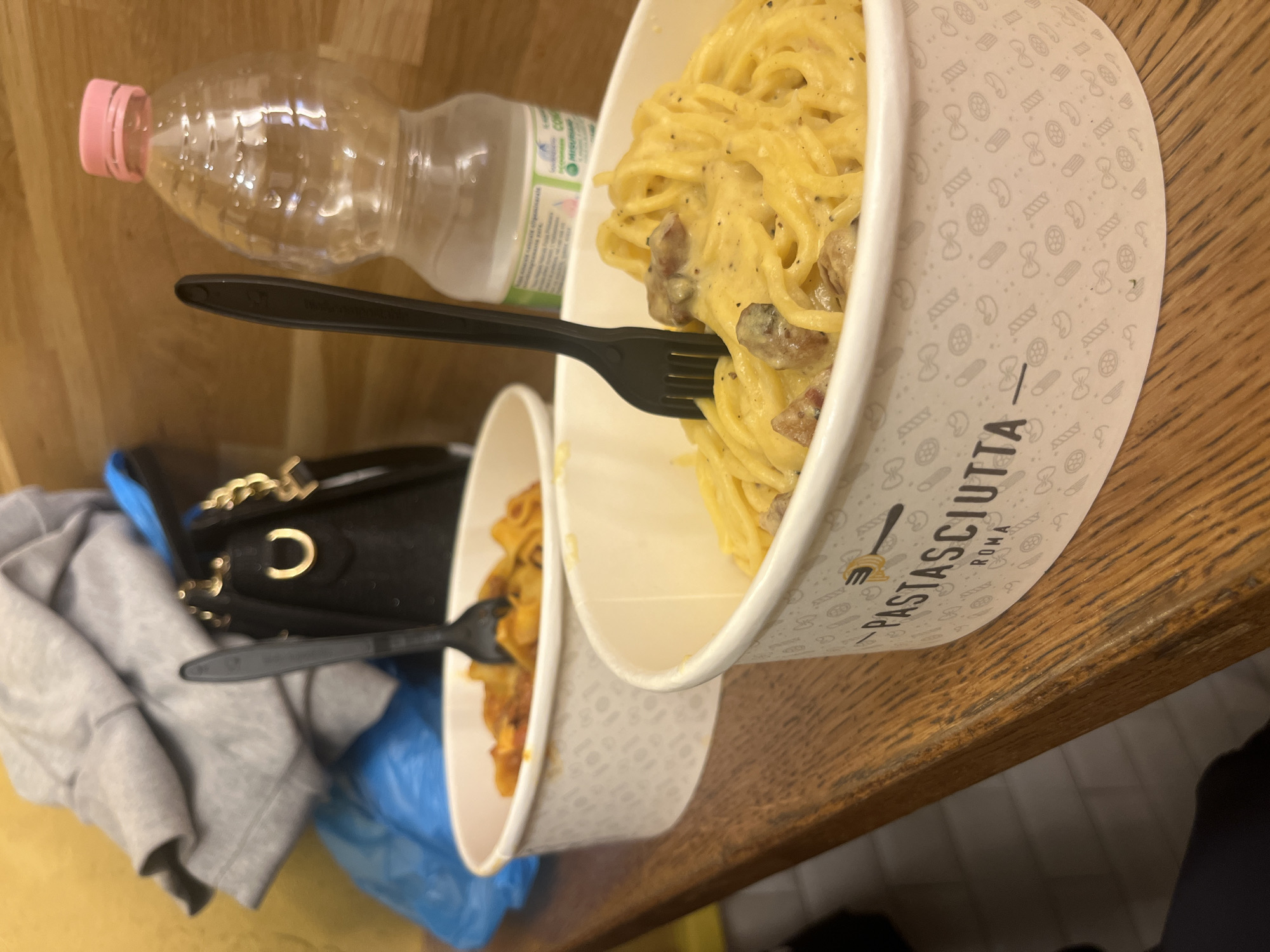 The image presents a close-up view of two takeaway pasta meals arranged on a light brown wooden table or counter.
In the foreground, a large white paper bowl is filled with a portion of pasta, likely spaghetti or bucatini, coated in a rich, creamy yellow sauce, characteristic of Carbonara. Visible pieces of browned meat, possibly guanciale or bacon, are mixed throughout the pasta. A black plastic fork is stuck into this dish. The exterior of the bowl is adorned with faint grey patterns of various pasta shapes, and the text "PASTASCIUTTA ROMA" is clearly visible in black lettering.
Behind and slightly to the left, a second, smaller white paper bowl contains another pasta dish, featuring pasta coated in a reddish-orange sauce, suggesting a different variety such as Amatriciana or Ragu. Another black plastic fork is present in this bowl.
To the left of the pasta bowls, a clear plastic water bottle with a pink cap is visible. Below the level of the table, partially obscured, are personal belongings: a black handbag with a gold chain strap, a grey folded hoodie, and what appears to be a blue plastic bag or object. The immediate background consists of a wooden wall.
The scene depicts a casual dining experience, likely at a takeaway establishment, with the "PASTASCIUTTA ROMA" branding explicitly indicating the meals are from Rome, Italy. The lighting suggests an indoor setting, but the time of day is not discernible.