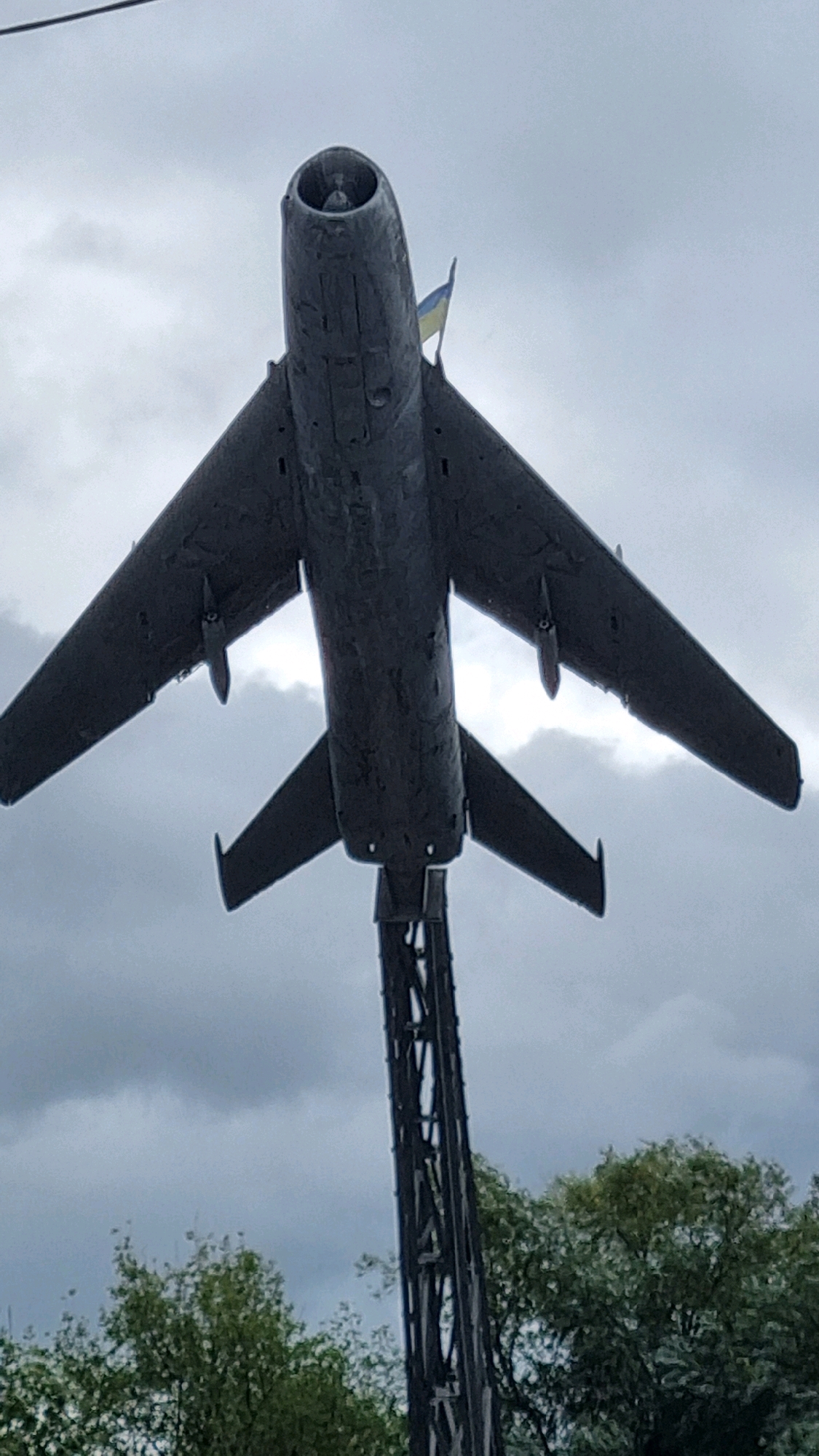 An underside view of a stationary, dark gray jet fighter aircraft is mounted on a tall, dark, lattice-like support structure. A small Ukrainian flag, with blue on top and yellow on the bottom, is affixed to the rear of the aircraft, just behind the cockpit. The aircraft is positioned against a backdrop of an overcast sky with heavy clouds. In the lower portion of the frame, the tops of green trees are visible. This monument is located in the city of Dubno, Ukraine.