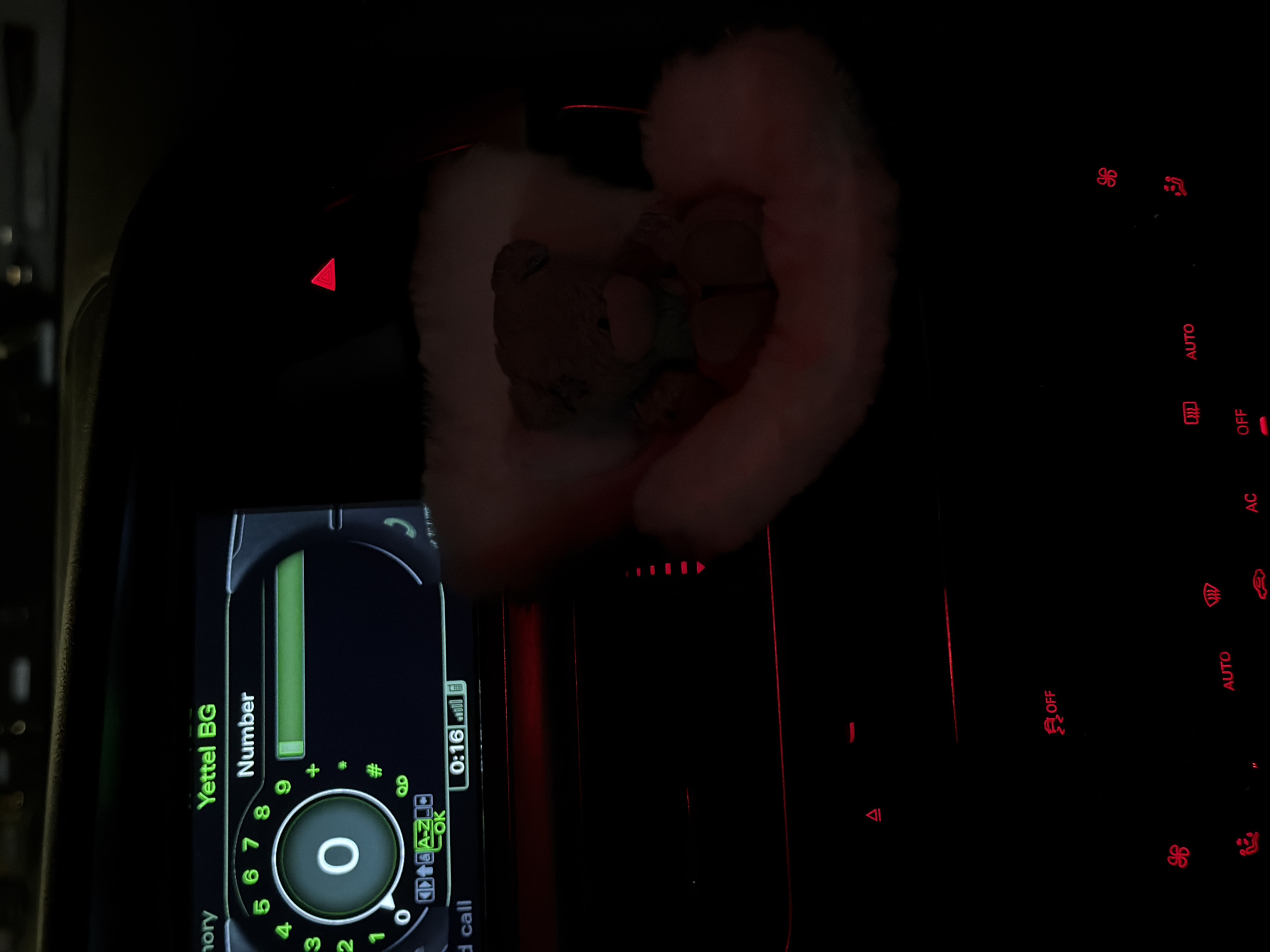 The image shows the illuminated dashboard of a car, partially obscured by a fluffy teddy bear in the center. The dashboard features illuminated climate control buttons to the right, and a screen displaying a car phone interface. The screen indicates a call in progress, with the network provider labeled "Yettel BG," and a call duration of 0:16. The location shown is in Bulgaria.