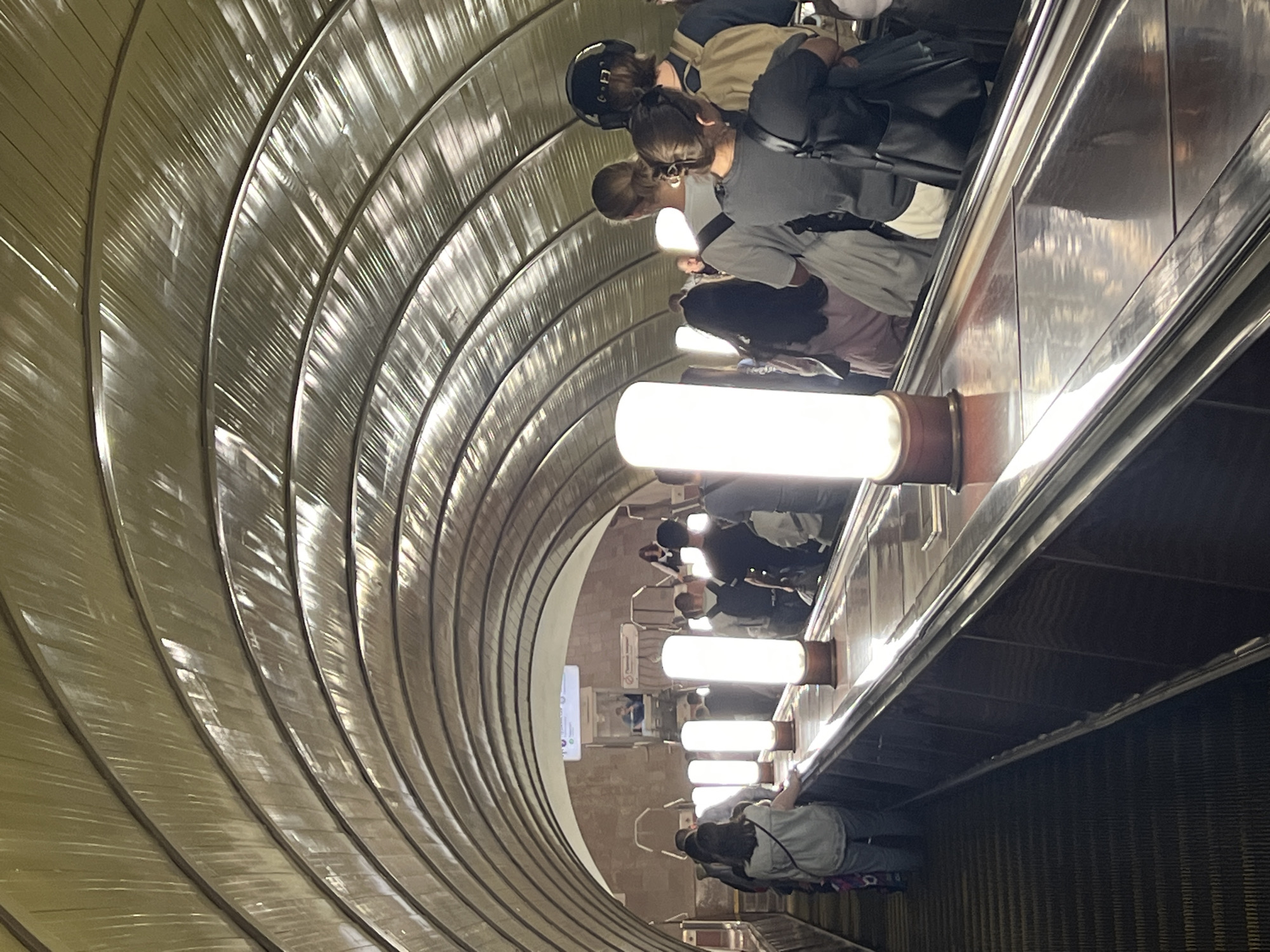 This media file shows a view of a curved escalator in a Moscow, Russia metro station. The escalator features rows of metallic siding and bright cylindrical lights. Several people are visible on the escalator, some moving up and others down. Most of them appear to be looking at their phones or engaging in conversation. They are dressed in casual attire, suggesting it's a normal day. The lighting is bright and artificial, typical of an underground metro environment. There are no clear indicators of the time of day or weather conditions. The scene evokes a sense of routine and everyday commuting. Some signage is visible in the background, but the text is too small and blurry to be deciphered.