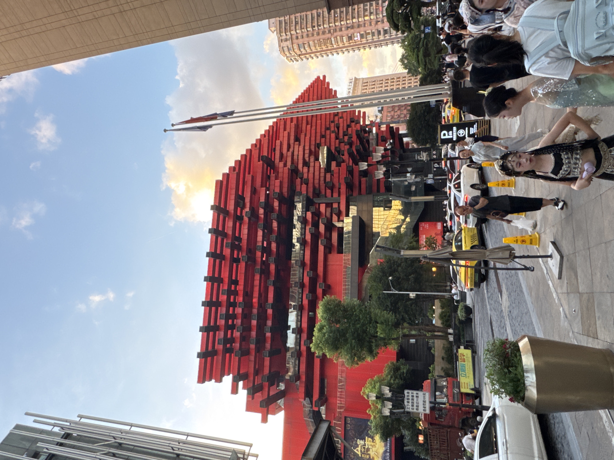 The image depicts a street scene in Yuzhong District, China, on a clear day with a mix of sun and clouds. A prominent, multi-layered red structure with black accents dominates the background, its design featuring horizontal stacked elements. To the right of this structure, two flags fly from poles. Adjacent to the red building, on the left, is a tall, modern glass-and-metal building. In the mid-ground, traffic flows on a street with cars, including yellow taxis, and a red truck. Pedestrians are visible on the sidewalk in the foreground and mid-ground. One young person in the foreground is dressed in a decorative costume with braided hair. Other individuals are dressed in casual clothing. A large, gold planter containing greenery is positioned on the sidewalk. Streetlights, traffic cones, and signs are also present. The overall impression is a bustling urban environment.