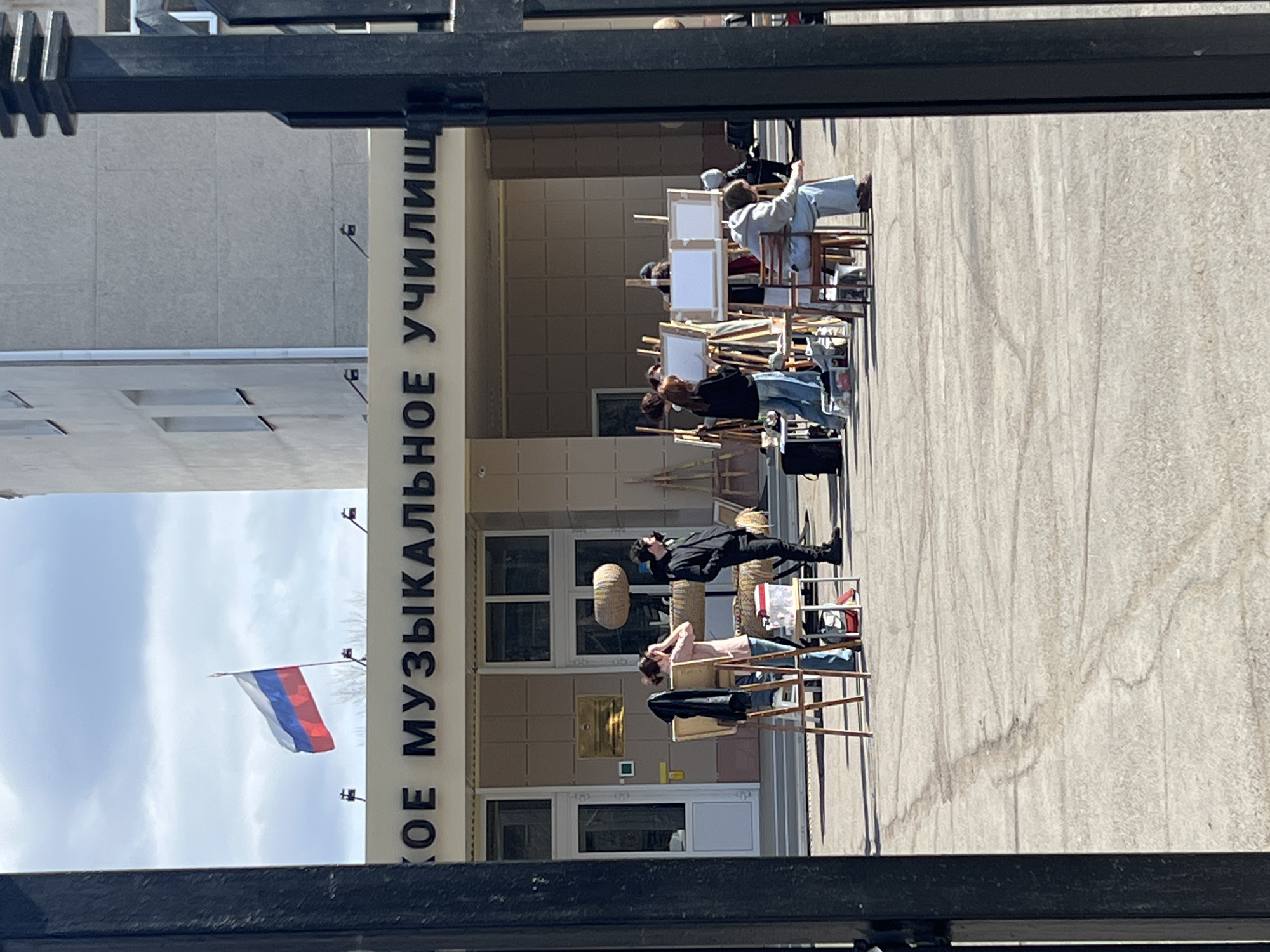 The image depicts a group of people painting outdoors in front of a building. Several individuals are seated on chairs, each with an easel and canvas set up before them. It appears they are engaged in an art class or workshop. The building's facade has signage in Russian, "ОЕ МУЗЫКАЛЬНОЕ УЧИЛИЩЕ," suggesting it is a music school. A Russian flag is visible on a pole attached to the building. The location is likely in the city of Chaikovsky, Russia, given the Russian signage and flag. The day seems bright and sunny. The scene is viewed through a metal fence or gate.