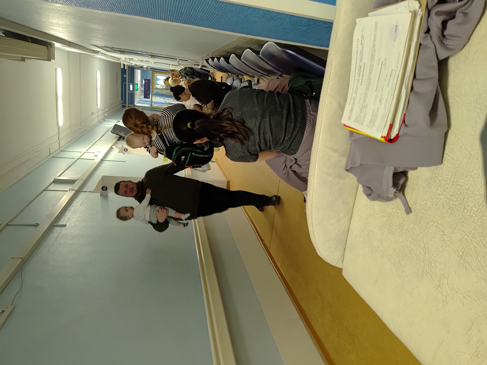 The image captures an indoor waiting area, likely within a medical facility or public building in Chaikovsky, Russia. The scene is brightly illuminated by overhead fluorescent lights, suggesting daytime conditions.
In the foreground, a bearded man, dressed in dark attire, holds a baby wearing light-colored clothing. Both are looking towards the viewer, with their reflections visible on the polished floor.
To the right, several individuals are seated in rows of blue-upholstered waiting chairs. A woman with long dark hair, wearing a dark top and light pants, leans forward in one of the chairs. Further back, another woman with brown hair and a striped shirt holds a fair-haired baby. Other adults are scattered throughout the seating area.
In the immediate foreground on the right, a light-colored couch or bench is visible. A grey sweatshirt is draped over it, with a stack of papers resting on top. Part of the visible text on one document, "Медицинский полис обязательного...", translates to "Mandatory medical insurance policy...", strongly indicating a medical setting.
Notable details include a green "EXIT" sign with a left-pointing arrow visible in the background, and a small sign displaying the number "252". The atmosphere appears calm, with individuals patiently waiting.