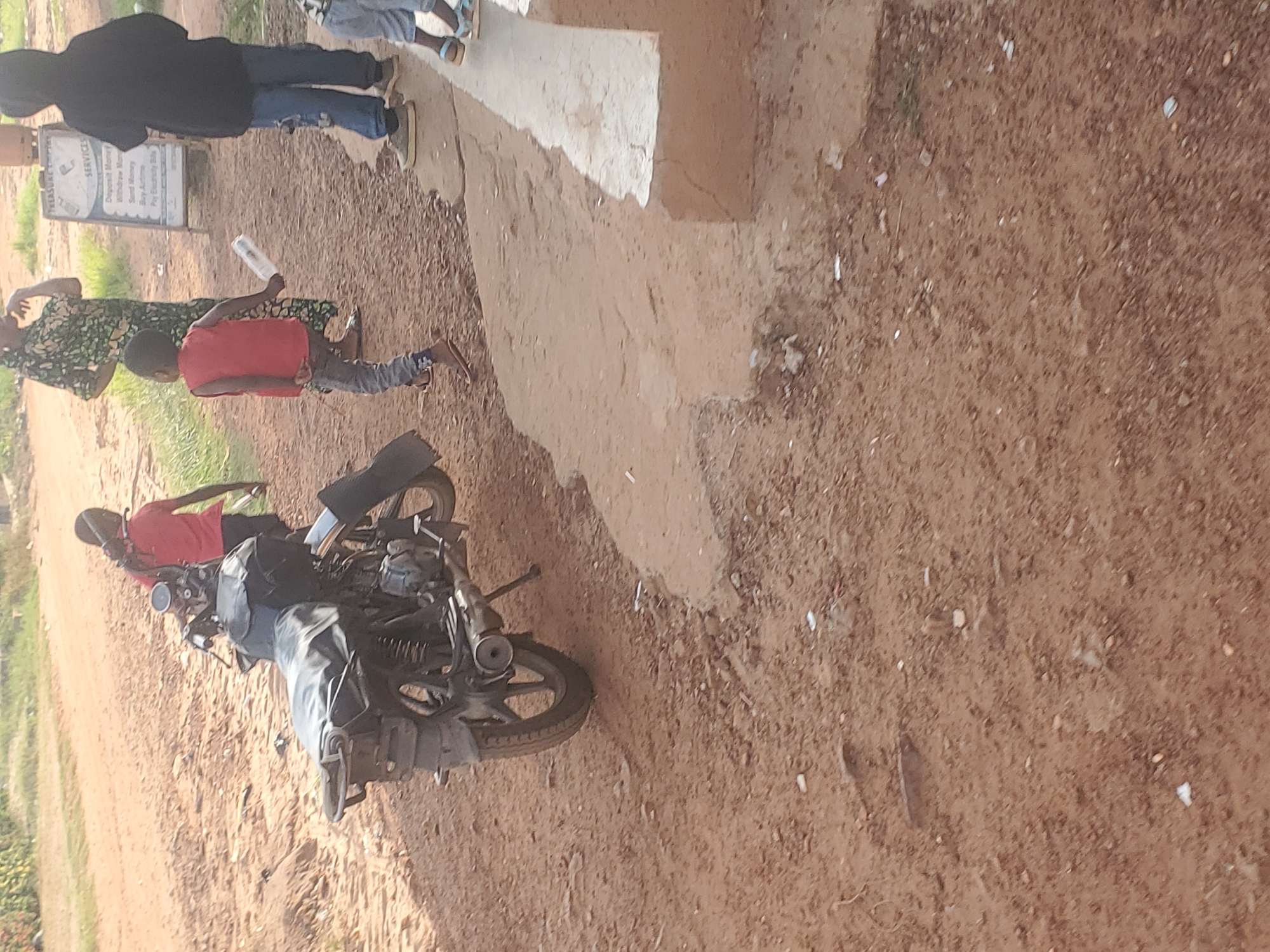 The image depicts a scene in Ukwu-Abwa, Nigeria, showing a group of people standing near a motorcycle on a dirt road. One individual is seated on the motorcycle, wearing a red shirt. Nearby, a woman in a patterned dress is standing next to a child in a red shirt who is holding a water bottle. Another person in a dark hoodie and jeans stands on a concrete platform. A sign with text is partially visible in the background. The ground is primarily dirt and reddish-brown.