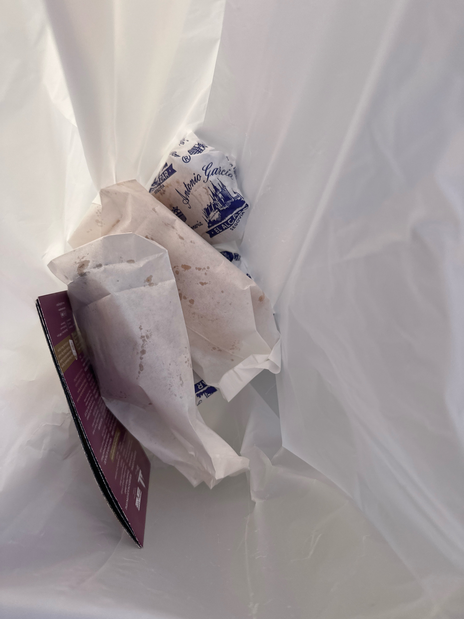In the city of La Lastrilla, Spain, a white plastic bag contains several wrapped food items. One item is wrapped in white paper with brown spots, and another is wrapped in paper bearing the blue inscription "Antonio Garcia" and "EL ALCAZAR SEGOVIA". A folded purple and black pamphlet is also visible in the bag, partially obscuring the food items. The bag is open, revealing its contents against a white background.
