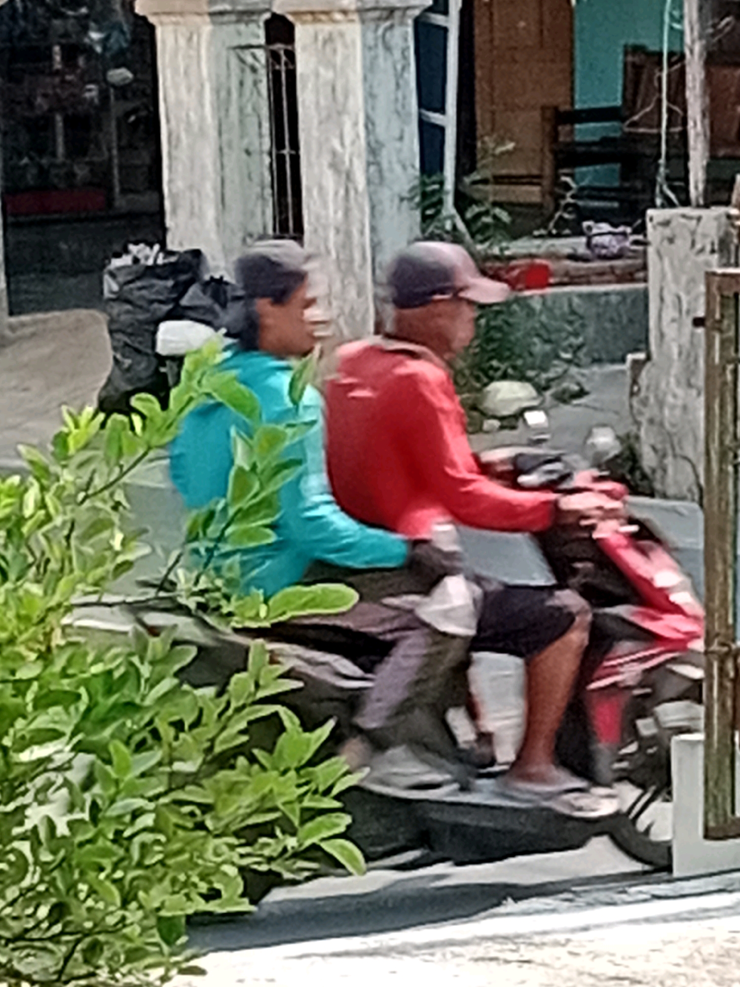 In Waru, Indonesia, two individuals are observed riding on a motorcycle. The person in the front, the driver, is wearing a red long-sleeved shirt and dark shorts. They have a dark baseball cap on. The passenger behind them is wearing a turquoise long-sleeved shirt and a dark baseball cap. The motorcycle is red and black. In the foreground, lush green foliage partially obstructs the view. Behind the motorcycle, a textured wall and part of a building are visible. The scene appears to be set on a street.