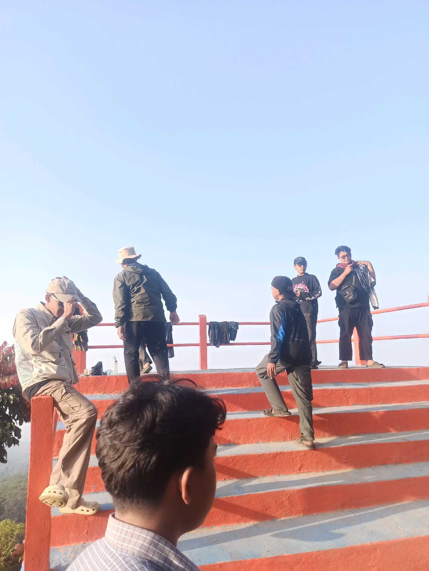 The image shows a group of people on an outdoor staircase with an orange railing under a clear, light blue sky. The scene appears to be a scenic overlook or viewpoint, possibly in Cileunyi, Indonesia.
In the foreground, a person's back is visible, with dark, curly hair and a checkered shirt. They are looking towards the group of people on the stairs.
On the stairs, there are at least five individuals. One person in the foreground of the stairs, on the left, is wearing a light-colored jacket, tan pants, and tan clogs. They are sitting on the railing and adjusting their hat. Further up the stairs, a person in a dark green jacket and dark pants stands facing away from the camera. To their right, a person wearing a black t-shirt, black pants, and a dark baseball cap is looking down. Next to them, another person in a black t-shirt and black pants is standing, holding a bag over their shoulder and a scarf around their neck. On the far right of the stairs, a person in a black jacket and dark pants is walking up.
Several articles of clothing are draped over the orange railing, including black t-shirts and pants. A white water bottle and a dark bag are also visible on the stairs.
The stairs themselves are wide and appear to be made of concrete, with orange painted risers and light blue or grey treads. Shadows from the railing and the people cast patterns on the stairs. The background shows a blurred landscape with some greenery, suggesting a natural or semi-natural environment. The overall lighting suggests it is daytime, likely morning or late afternoon due to the soft light.