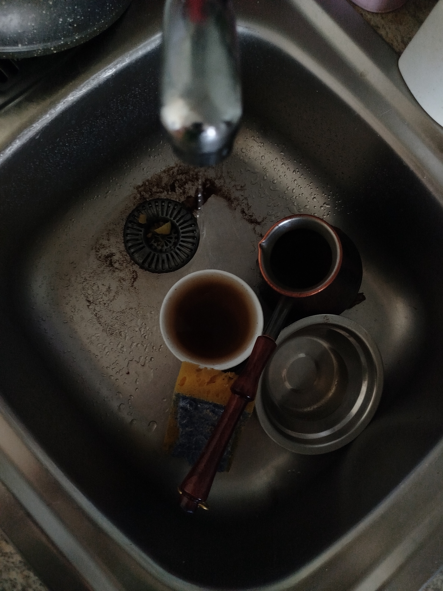 An overhead view captures a stainless steel kitchen sink filled with water and some residue, suggesting recent use. A chrome faucet is visible at the top, partially out of focus. In the sink are several items: a dark-colored cezve containing a dark liquid, possibly coffee; a white cup with a similar dark liquid; a yellow and blue sponge; and a silver-colored metal cap or lid. A drain with a black grate is also present. The overall scene is indicative of a sink preparing to be cleaned or in the process of being cleaned. This image does not provide any specific geographical markers that would allow for precise location identification within the city of Perm, Russia.