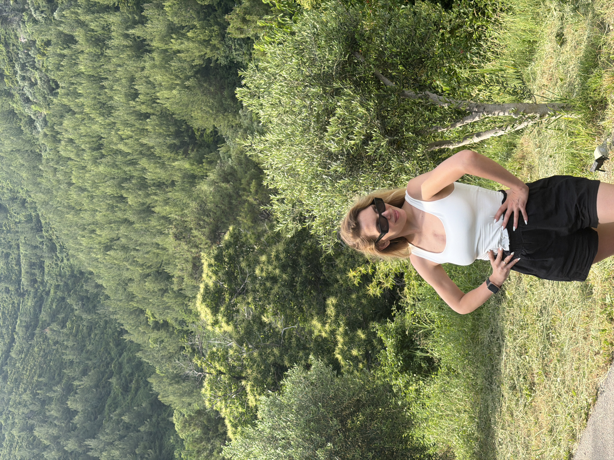 A woman stands near a mountain covered with greenery. She wears sunglasses, a white tank top, and black shorts. Her hands are on her hips. The location is Budva, Montenegro.