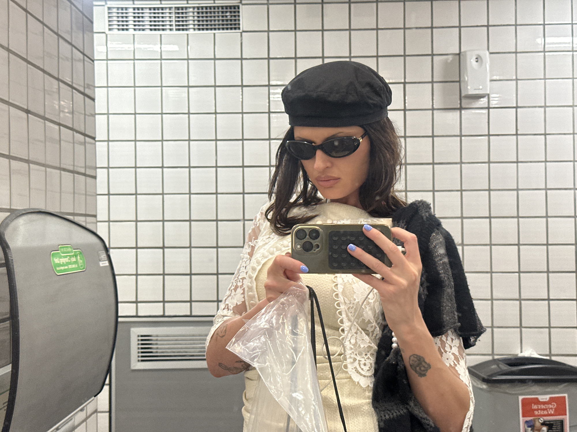 The image shows a woman taking a selfie in a bathroom. The bathroom features white square tiles on the wall, a metal hand towel dispenser on the left, and a white air freshener dispenser on the wall behind her. Below the towel dispenser is a ventilation grill set into the wall. To the right, a trash receptacle is visible. The woman is wearing a black beret and small sunglasses. She is holding a smartphone with a green case to take the photo. In her other hand, she has a clear plastic bag. She is wearing a white dress with lace detailing and a dark, possibly knitted, garment is draped over her shoulder. The location cannot be determined from the image.