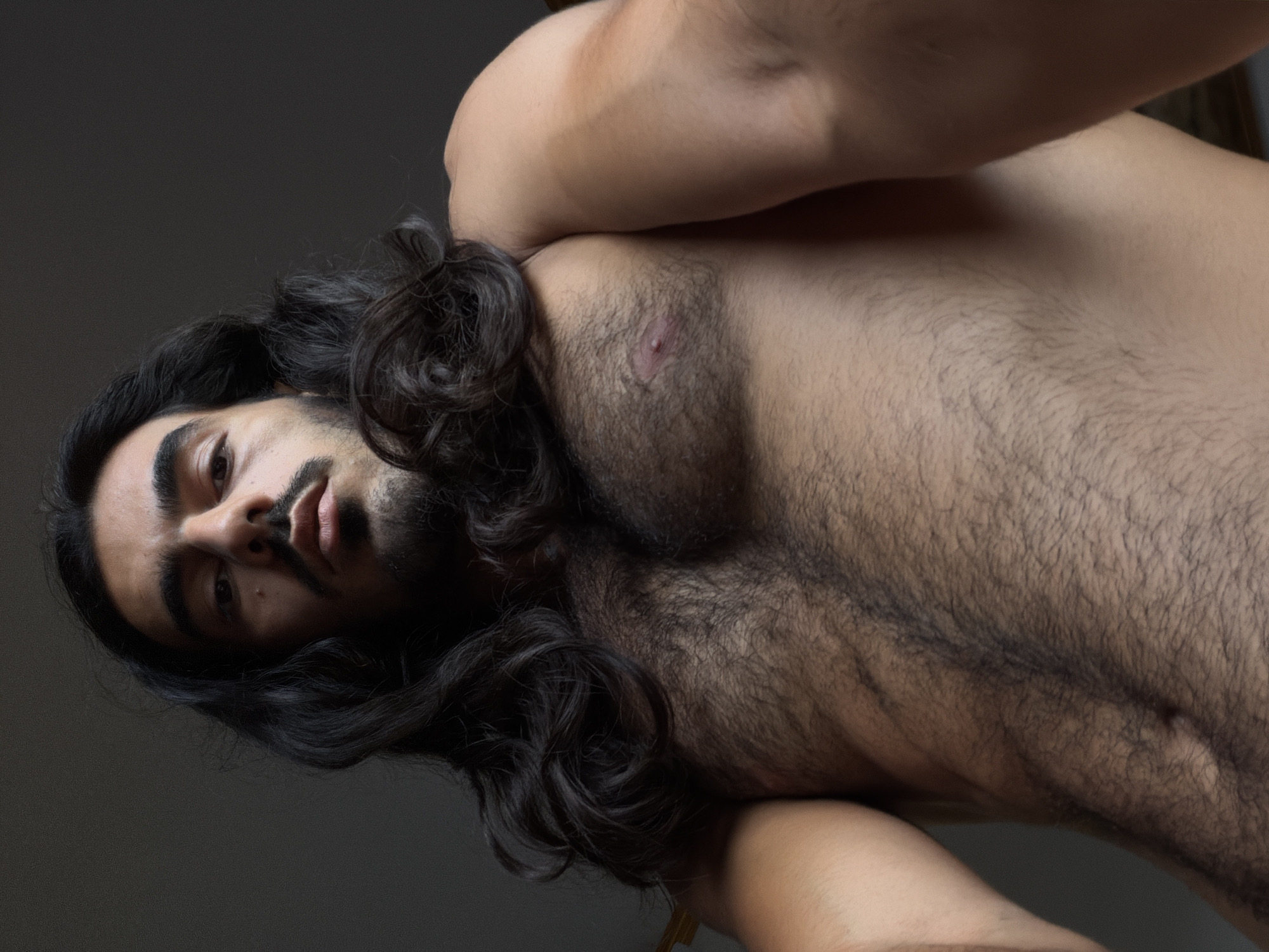 A shirtless man with long, dark, wavy hair and a full beard is featured in a low-angle, close-up shot. He is lying down with his head tilted, gazing directly into the camera with dark, intense eyes. His right arm is bent and raised, resting behind his head, while his left arm is also bent, positioned to his side or beneath him. His chest and upper abdomen are prominently displayed, covered with dense dark hair, with one nipple visible and appearing somewhat reddish. The man has a warm skin tone and a small mole visible on his left cheek.
The background is a plain, dark gray, suggesting an indoor setting with soft, diffused lighting that highlights his features and the texture of his hair. His expression is neutral, bordering on contemplative or serious. There are no visible location cues within the image itself, though the provided context is North Kuta, Indonesia. No text is visible in the media file. The image's composition emphasizes his direct gaze and physical form.