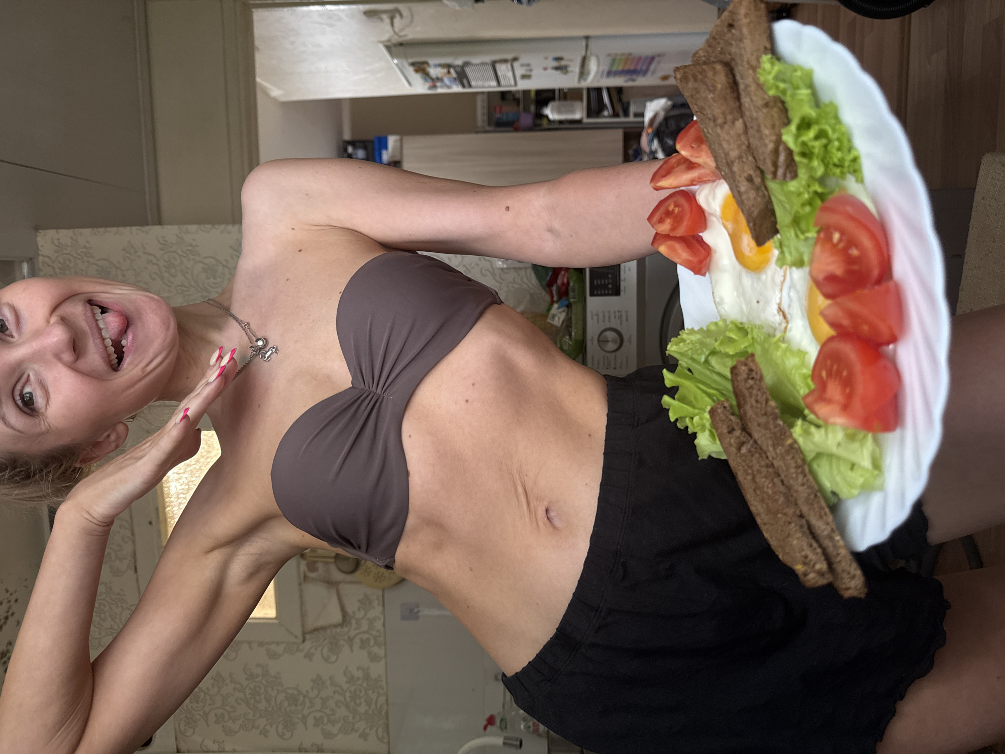 A woman with blonde hair and fair skin is holding a plate of food. The plate contains fried eggs, tomatoes, lettuce, and dark bread. She is wearing a brown bra and black shorts. She is smiling and sticking her tongue out. A silver necklace with a charm is visible around her neck. She has long, pink fingernails.
The background shows a cluttered kitchen with a wallpapered wall and various appliances. Based on the available visual information, it's impossible to confirm the location in the city of Chaikovsky, Russia.