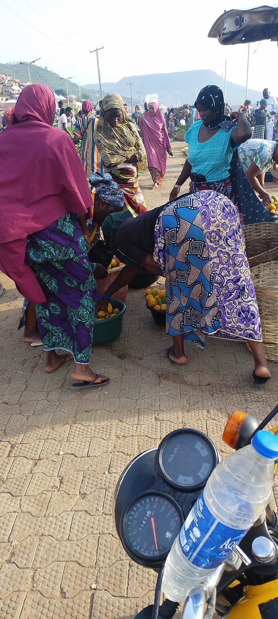 Here is a description of the image:
The image shows an outdoor scene in Lokoja, Nigeria, possibly a marketplace. Several women, likely vendors or shoppers, are gathered around containers filled with yellow fruits, possibly oranges or tangerines. The women are dressed in traditional African clothing with colorful patterns. They are picking through the fruit, suggesting a purchasing transaction or sorting process.
In the foreground, the dashboard of a Bajaj motorcycle is visible, including the speedometer which reads 11473 km. A water bottle is also placed on the motorcycle. The ground is paved with patterned concrete blocks. In the background, more people are visible, along with buildings and a hill in the distance, confirming that the image was taken within an urban area. Street lighting is also apparent.