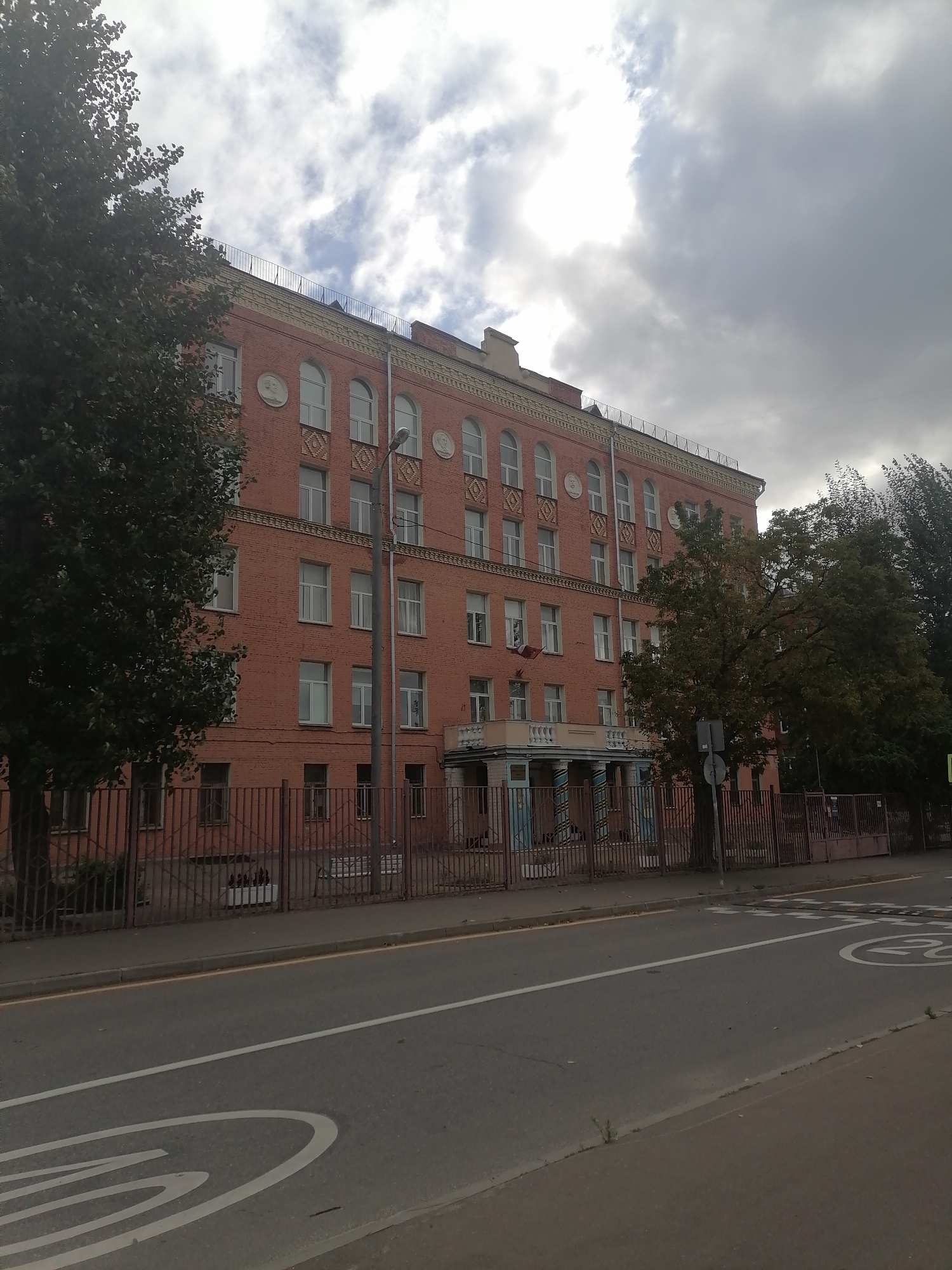 The image shows a brick building with many windows, typical of an educational institution, in Moscow, Russia. The building has an ornate facade with decorative elements and a small balcony above the entrance. In front of the building, there is a metal fence and a street with road markings. Trees are visible on both sides of the building. The sky is cloudy and overcast.