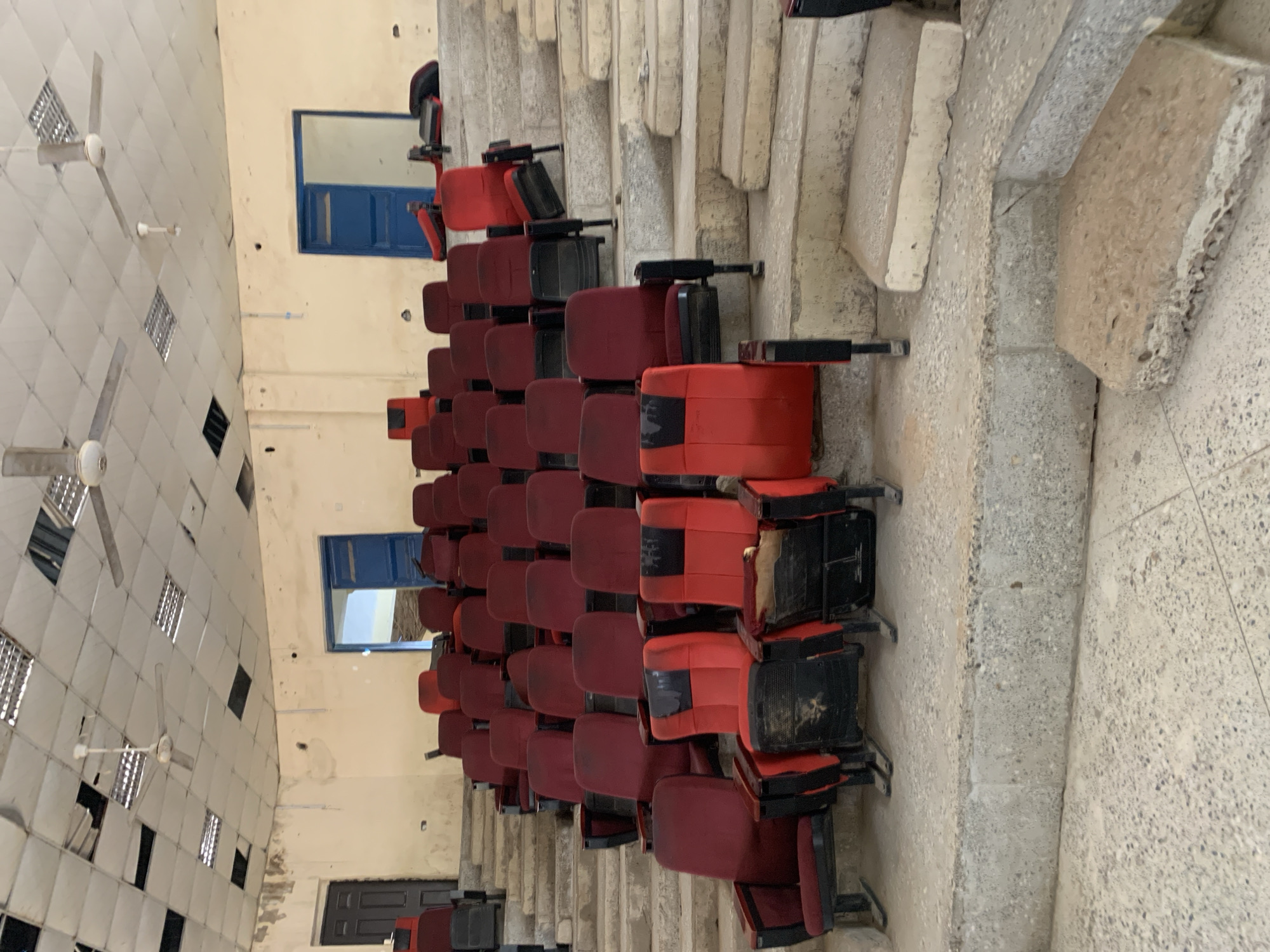 This media file depicts an indoor scene, likely an auditorium or lecture hall, in Jalingo, Nigeria. The room features rows of red, stadium-style seating, most of which appear empty. Some seats are flipped up, revealing black undersides, while others are down. The seating is arranged on a tiered structure made of concrete.
The ceiling is visible on the left side of the image, with a white tiled pattern and several large, white ceiling fans. Some of the ceiling tiles are missing or damaged, revealing dark openings. Fluorescent light fixtures are also present in the ceiling.
Further back in the room, two windows with bright blue frames are visible on a cream-colored wall. Below one of the windows, a dark door can be seen. The overall impression is of a somewhat dated or possibly under-maintained public space. There is no visible text within the image. The lighting suggests it is daytime, but there are no clear indicators of the weather or a specific event taking place. The scene is quiet and unoccupied.