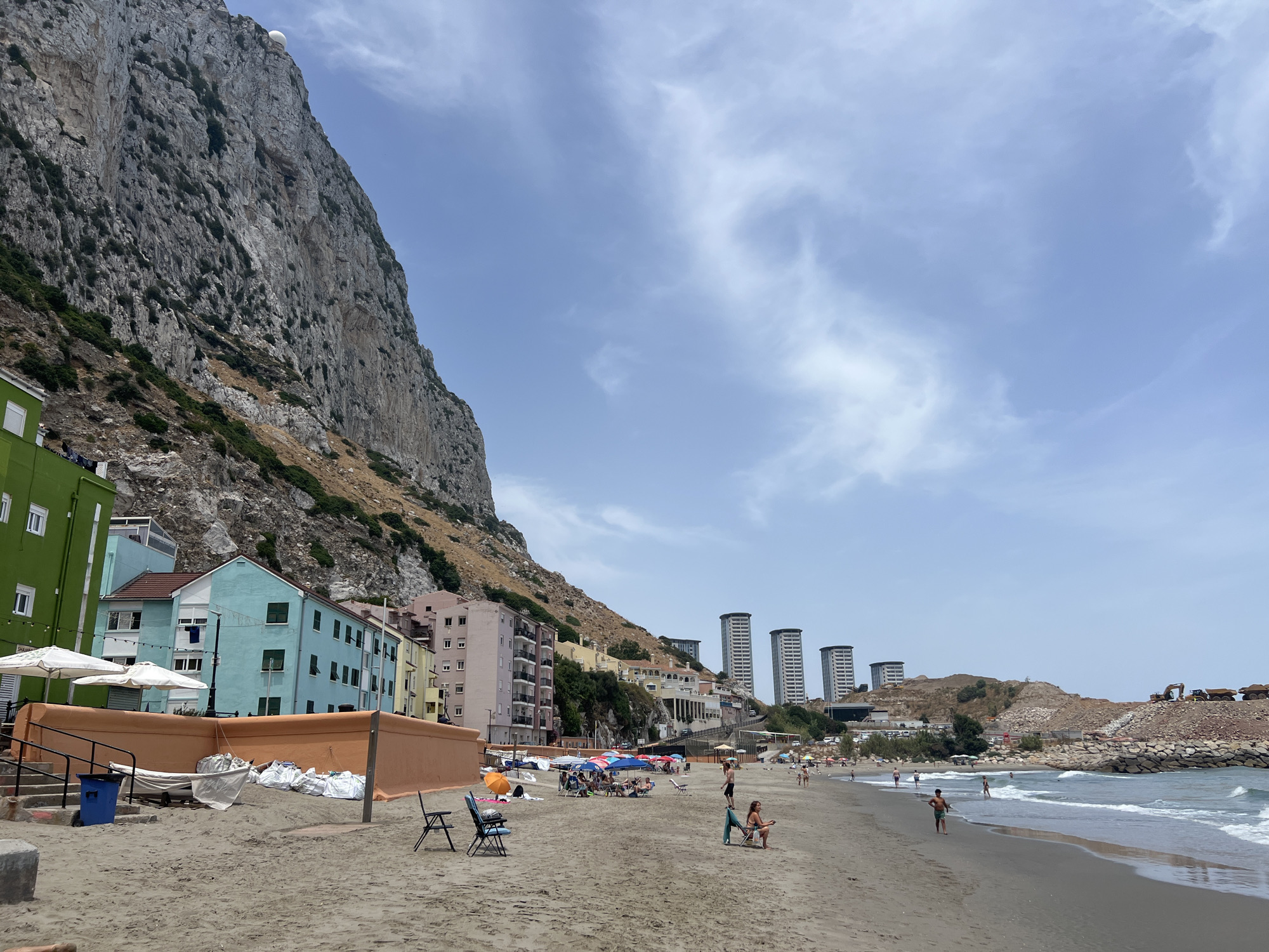 This image depicts a sunny day at a beach in Gibraltar. The scene is dominated by the imposing Rock of Gibraltar on the left, its rocky face covered in sparse vegetation. Below the rock are colorful buildings, ranging in hue from bright green to pale yellow, with several apartment blocks situated further back and higher up.
The beach itself is sandy, with a calm sea lapping at the shore. Numerous beach umbrellas are scattered across the sand, providing shade for people who are relaxing or swimming. Several individuals are visible on the beach, some sitting in chairs, others walking or playing. Some are wading in the shallow water. The atmosphere appears relaxed and leisurely, indicative of a pleasant day out.
In the distance, several modern high-rise buildings stand out against the skyline. To the far right, there is evidence of construction or land reclamation, with heavy machinery like excavators and dump trucks visible on a rocky embankment. The sky is a light blue with scattered white clouds, suggesting fair weather and likely midday to early afternoon. No visible text is present in the image.