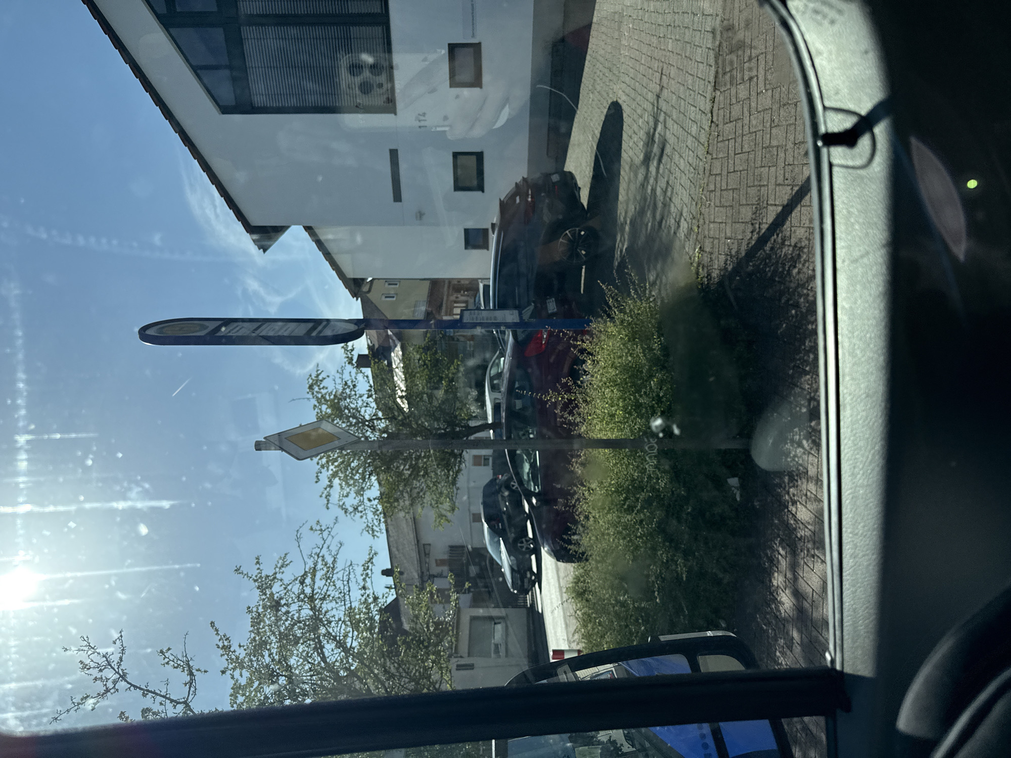The image captures a street scene in Volpertshausen, Germany, on a bright, sunny day. The perspective is from inside a vehicle, looking out through the windshield and side window. In the foreground, the interior of the vehicle is visible, including a dark-colored dashboard and a portion of the driver's side door.
Outside the vehicle, a paved street or sidewalk with a brick pattern is visible. Several cars are parked or driving on the street. A dark-colored SUV is parked in front of a white building with a red-tiled roof. To the left of the SUV, a red hatchback is also parked. Further down the street, other vehicles and buildings can be seen.
A prominent bus stop sign with a blue pole stands near the parked cars. A yellow diamond-shaped road sign is also visible, mounted on a pole behind a cluster of green bushes. The sun is shining brightly, casting shadows on the ground and creating lens flare in the image, particularly in the upper left quadrant. Trees with green foliage are present on the sides of the street, and the sky is a clear blue. The white building in the background features large windows with blinds and the number "114" is visible on its facade.