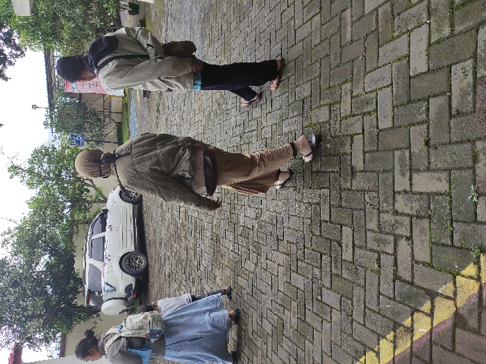 The image captures an outdoor scene in Kepanjen, Indonesia, featuring several individuals on a paved area during what appears to be daytime. The ground is covered with grey interlocking bricks laid in a herringbone pattern, showing some signs of wear and minor vegetation growth.

In the foreground, on the left, a person wearing a light blue long dress or skirt carries a colorful patterned backpack. Next to them, another individual is partially visible. Further back, two more people are seen walking or standing. One wears a brown jacket, light brown pants, and a helmet on their head, carrying a small bag. In front of them, another person is dressed in a light-colored jacket with dark accents, dark pants, and also carries a backpack. All visible individuals are seen from behind or the side, making their expressions undiscernible. Most are wearing sandals or open-toed footwear.

A white SUV is parked in the background on the left. The background also includes sparse trees and glimpses of buildings. A blue sign with a white capital letter "P," indicating parking, is visible through the foliage. A yellow painted line is present along the right edge of the paved area. The overall scene suggests a casual outdoor environment, possibly a pathway or parking lot area.