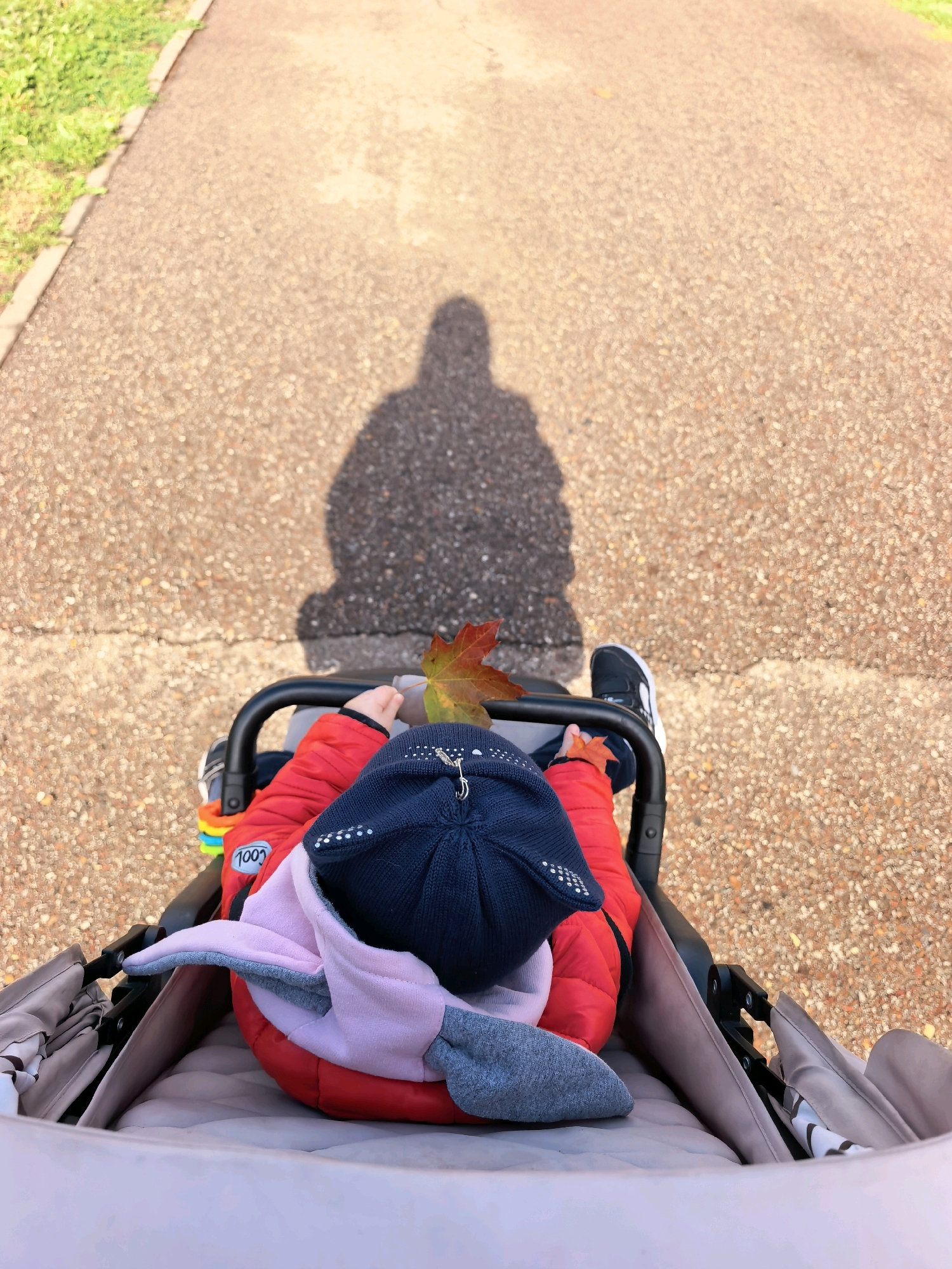A high-angle, medium shot shows a baby in a stroller, facing away from the camera. The baby is wearing a red puffer jacket, a dark blue knit hat with decorative rhinestones, and pink and gray ear-shaped accessories attached to the hat. A single red and yellow autumn leaf rests on the baby's jacket. The stroller is dark gray and has a patterned fabric lining. The baby's feet are visible, wearing black sneakers with white soles and laces. A colorful toy is attached to the stroller's handle. The ground is a textured, light brown asphalt path, with a strip of green grass and a concrete curb on the left side of the image. The shadow of a person, presumably the one taking the photo, is cast on the path in front of the stroller. The scene is outdoors on a sunny day, with the lighting creating a shadow that suggests it is late afternoon or early morning. No specific landmarks of Sterlitamak, Russia, are identifiable in the image; it depicts a generic park or pathway setting.
