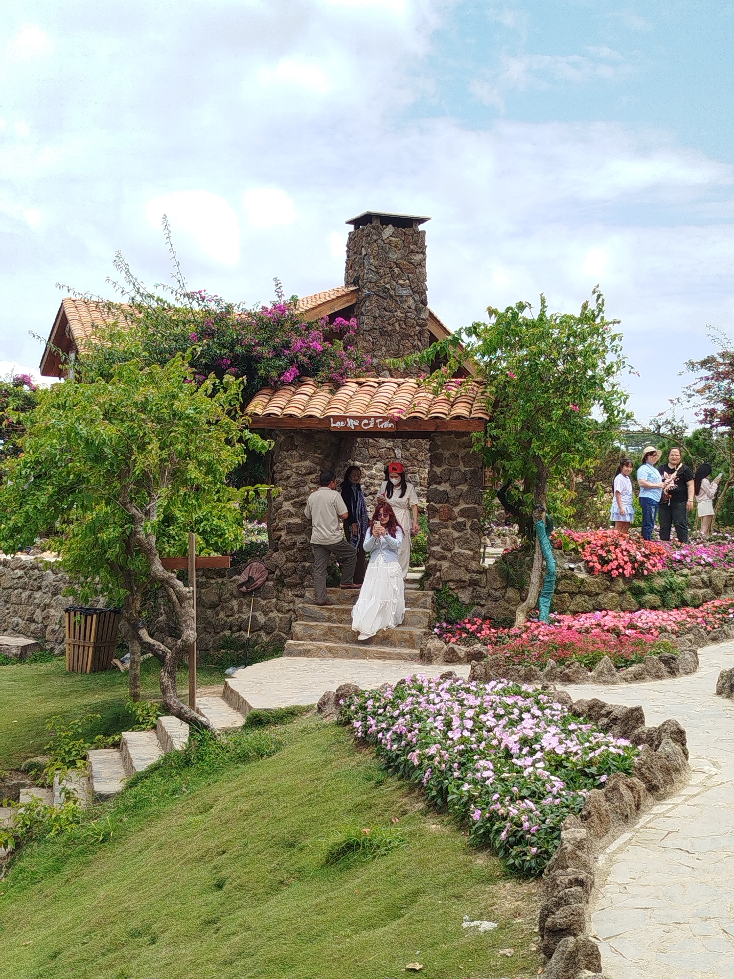 A stone house with a terracotta tile roof is situated in a garden setting. The house has a prominent stone chimney. Lush green trees and vibrant pink and purple flowers surround the structure, which appears to be a tourist attraction. A stone pathway winds through the garden, leading to the entrance of the house. Several people are present, some standing on the steps of the house, while others are scattered throughout the garden area, admiring the scenery. One woman in a long white dress is positioned on the steps, possibly posing for a photograph. Another woman, wearing a red face mask, stands behind her. To the right, a group of women are gathered near flower beds. The overall impression is a picturesque outdoor location, likely a cafe or attraction, in Xuân Thành, Vietnam, characterized by its natural beauty and architectural elements.