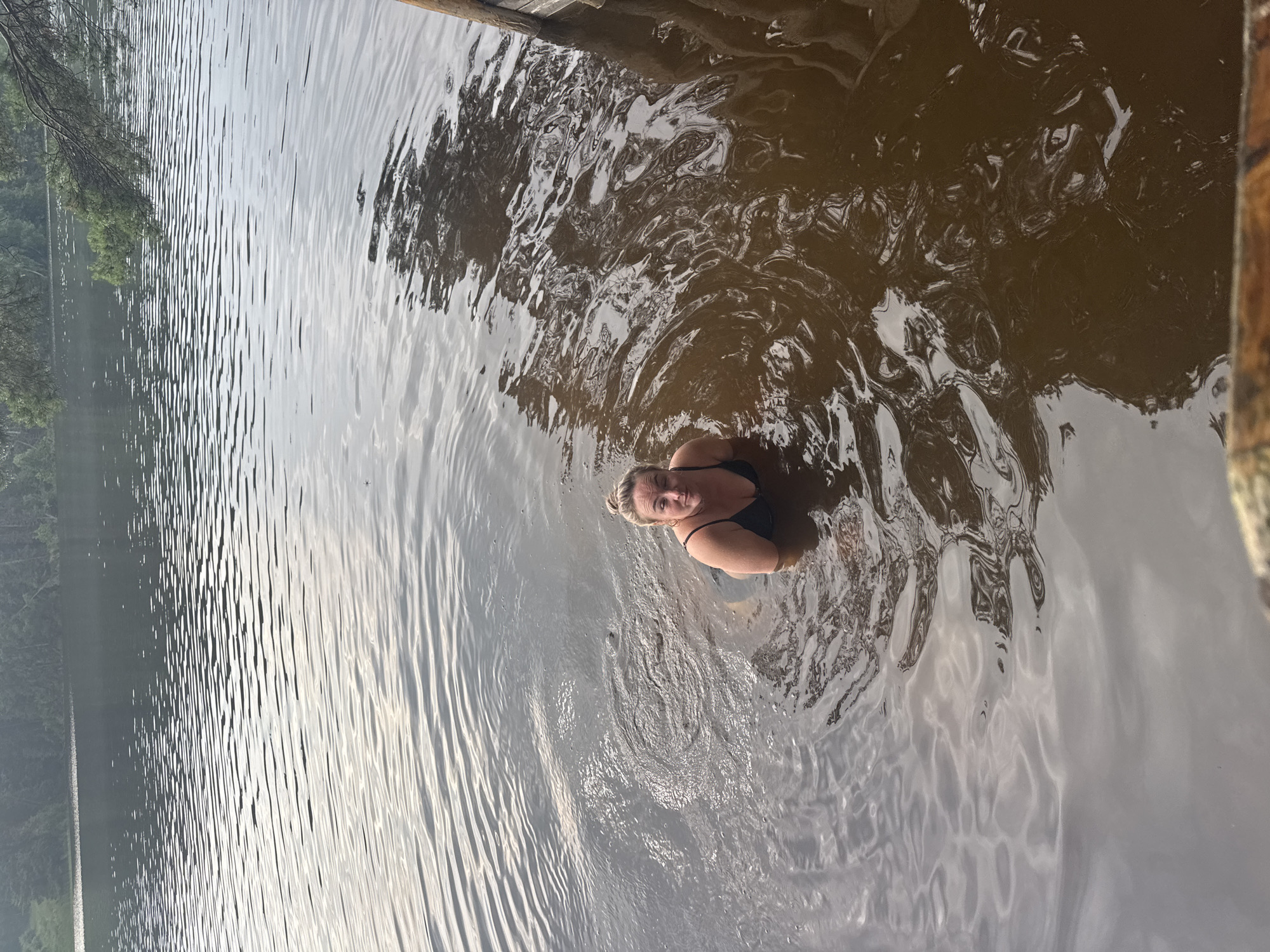 The image shows a person submerged in murky water, with their upper body visible. They are looking at the camera and appear to be smiling. The water is dark brown and appears to be a lake or river. There is a wooden structure to the right of the frame. The background consists of trees and a body of water. The location is the city of Pozdyshki, Russia.