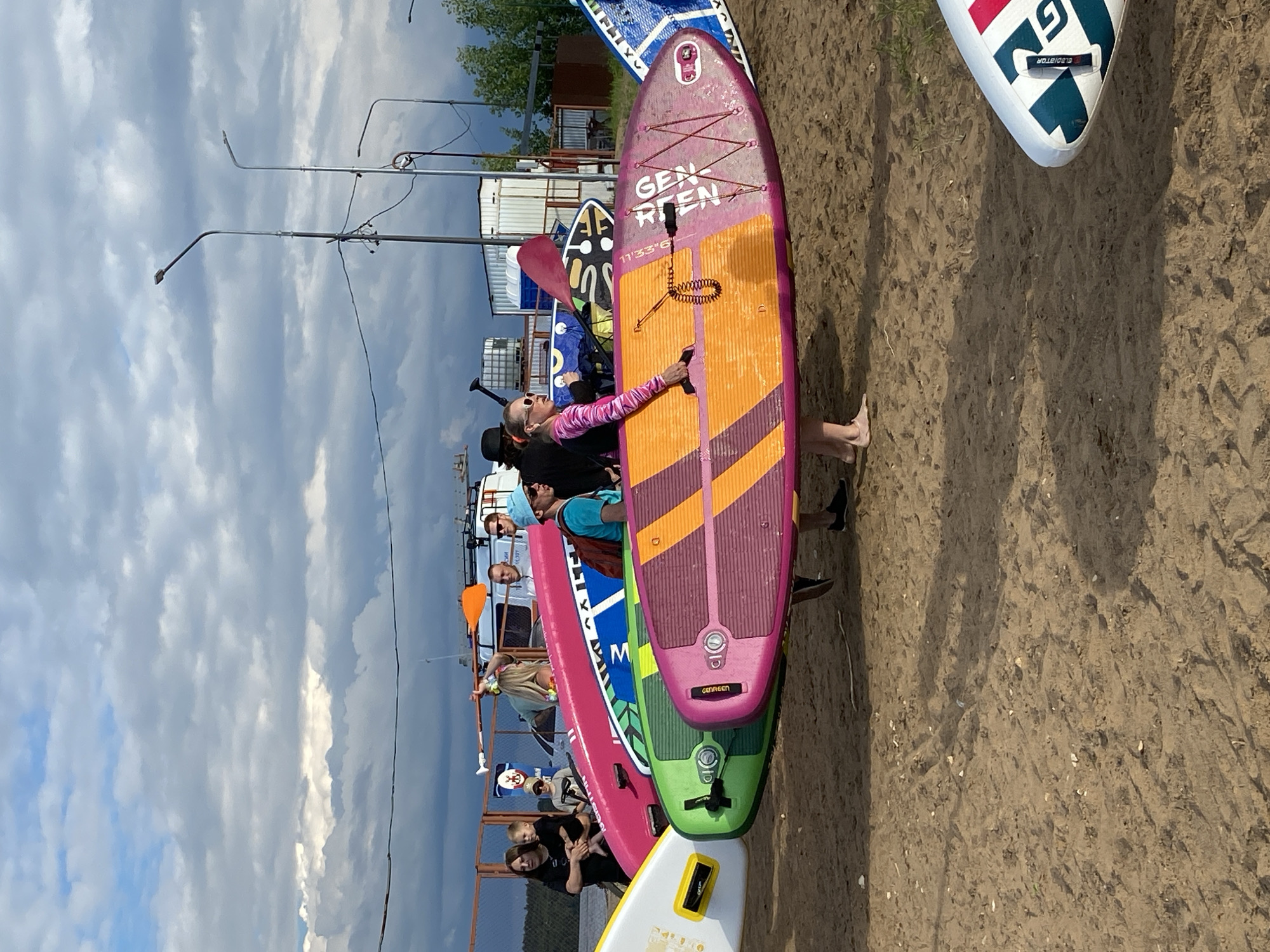 The image depicts a sandy beach scene in Chaikovsky, Russia, with a group of people preparing for or returning from a water activity. Several stand-up paddleboards (SUPs) are prominently displayed, with one pink and orange SUP in the foreground being carried by a woman. Other SUPs, in various colors including green and white, are also visible. In the background, there are more people, some carrying paddles, and structures that could be changing rooms or storage units. The sky is partly cloudy with a mix of sun and shade. The overall impression is of a recreational gathering by the water, possibly a SUP rental or club.