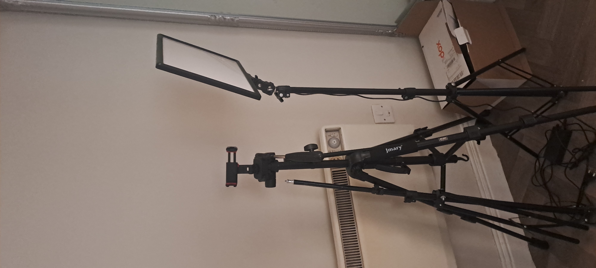 The image shows two black tripods set up in front of a white wall and a cream-colored radiator. The tripod on the left has a black phone holder at the top. The tripod on the right supports a rectangular white LED light panel. Electrical cords are visible on the floor, connecting to a black power adapter near the base of the left tripod. A cardboard box with red and white lettering is placed on the floor to the right of the right tripod, with some white objects protruding from it. The scene appears to be an indoor setting, possibly a room in Castlebar, Ireland, given the context of the request. There are no people present in the image. The lighting is even, suggesting the LED panel might be on or the room is well-lit. The floor is a dark wood or laminate material. A light switch and an electrical outlet are visible on the wall behind the radiator. A portion of a large, ornate mirror is visible on the far right edge of the image.