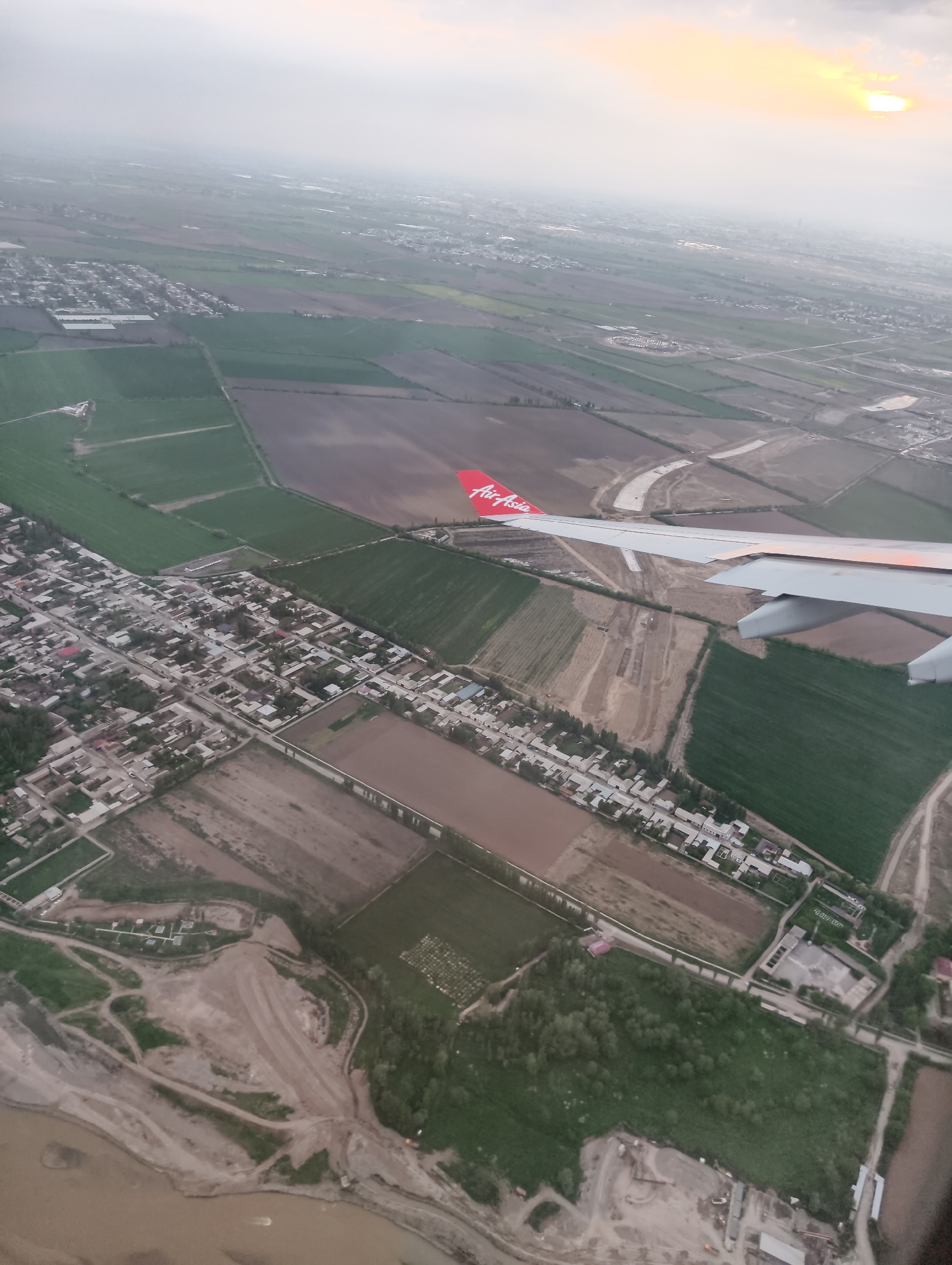 The image shows an aerial view from an AirAsia plane, looking down on a landscape with fields, a town, and a river. The sky is overcast with the sun visible near the horizon. The plane's wing and the AirAsia logo are visible in the foreground. The landscape features a patchwork of green and brown fields, a densely packed town with light-colored buildings, and a river with a muddy bank. Construction or industrial activity appears to be present near the river.