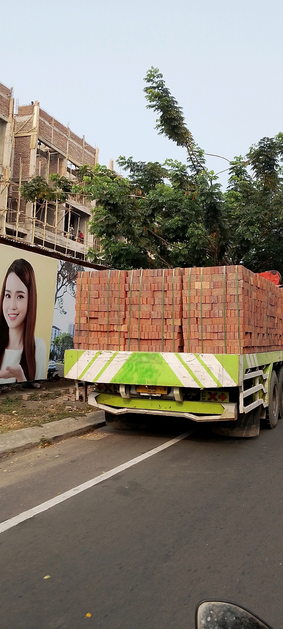 A flatbed truck carrying a large stack of bricks is traveling on a road in Gunung Putri, Indonesia. The truck is decorated with green and white diagonal stripes on its rear. To the left of the truck is a billboard featuring a woman with dark hair and a white shirt. In the background, a multi-story building is under construction, with scaffolding and workers visible. Lush green trees are interspersed with the building and the sky. The road has a white line marking its edge.