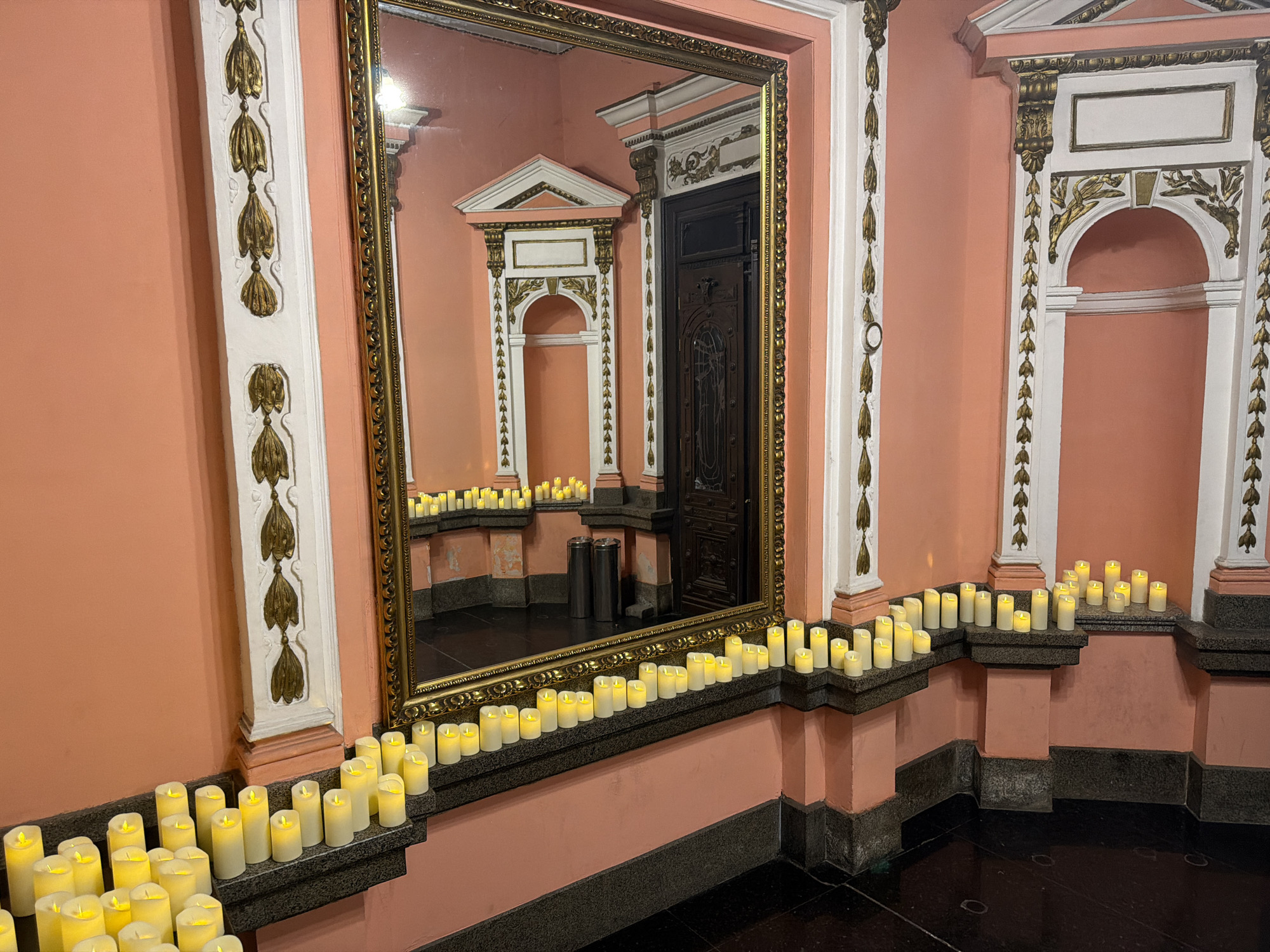 The image shows an interior space in Sofia, Bulgaria, featuring a large ornate mirror reflecting a replica of the setting. The walls are painted a light peach color and adorned with white architectural features highlighted with gold accents. LED candles are placed along the base of the wall and on ledges, creating a warm glow. The reflected image in the mirror shows a similar setting, including a dark wooden door and additional rows of LED candles. The floor is a shiny, dark material, likely marble or a similar stone. Two metal trash cans are partially visible in the reflection. No people are present in the image. The overall impression is that of a decorative and elegant space, perhaps a historical building or museum.