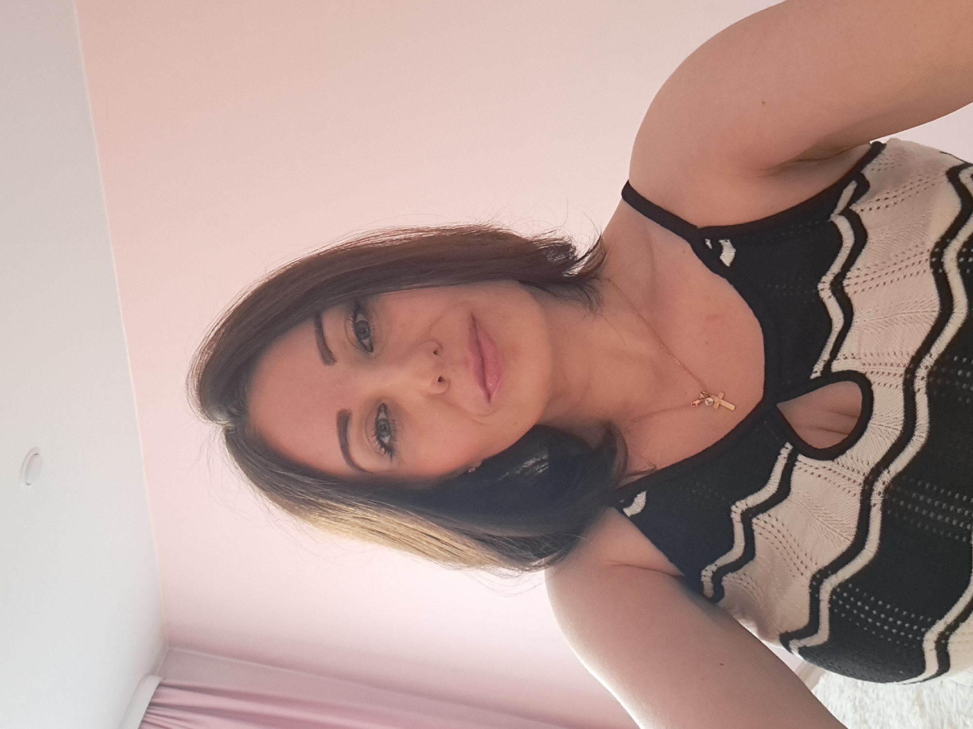 A woman with shoulder-length brown hair and blue eyes is looking at the camera. She is wearing a black and white striped top with a keyhole cutout and a gold necklace with a cross pendant. The background is a pale pink wall with a pink curtain on the left. There is a ceiling light visible at the top of the image. The overall setting suggests an indoor environment, likely a bedroom, in Irkutsk, Russia.