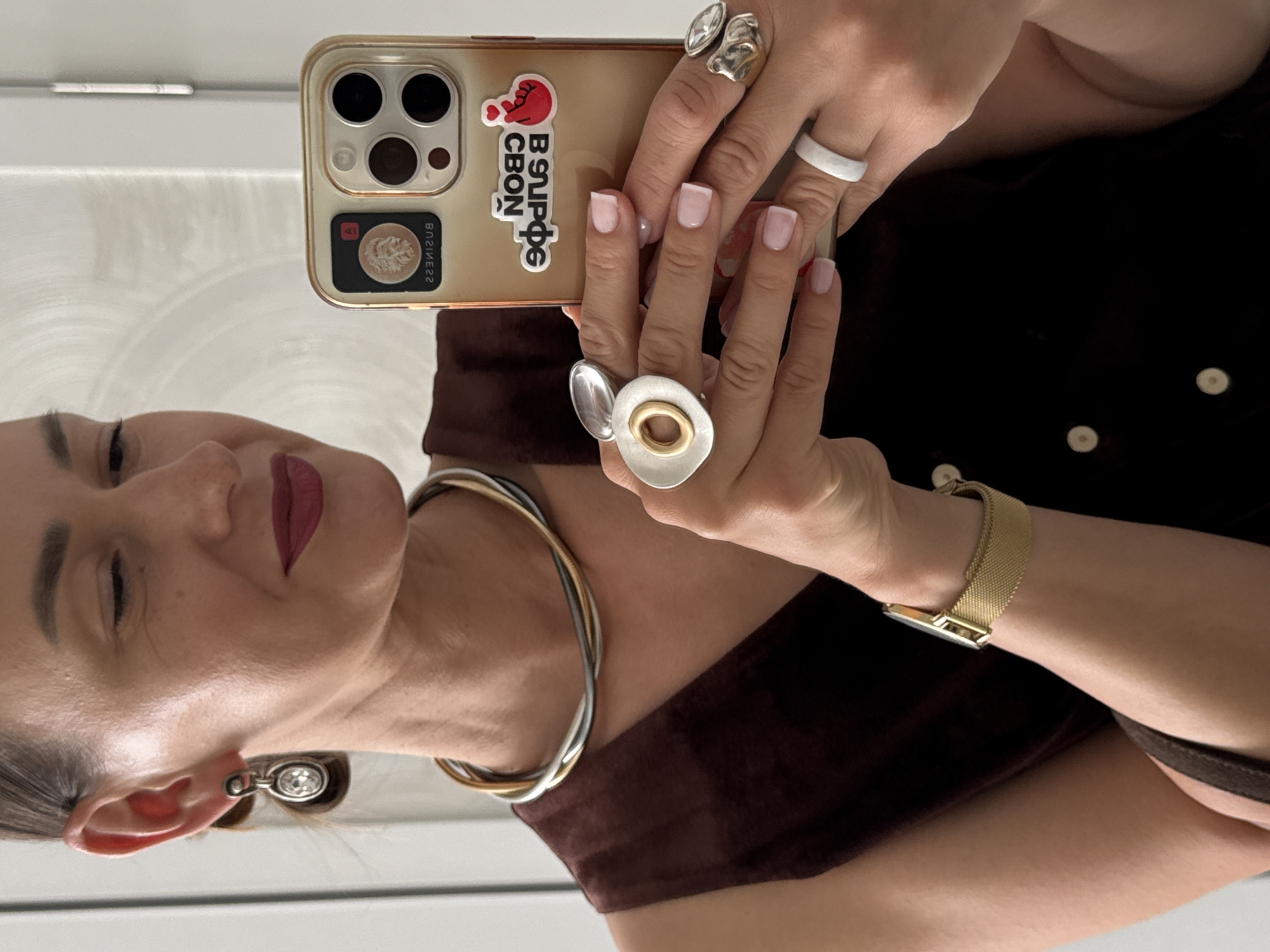A medium shot photograph captures a woman in what appears to be an indoor setting, possibly a residence, as indicated by the reflective surface of a door or mirror on the left and a plain wall on the right. The woman is looking into a smartphone, which she is holding with her right hand. She is wearing a brown sleeveless top, a multi-strand silver and gold necklace, a gold watch on her left wrist, and several rings on both hands. Her right earlobe features a dangling earring with a clear stone. The smartphone case is gold and has stickers on it. The background is out of focus, making it difficult to discern specific environmental details within the city of Sochi, Russia. The lighting suggests artificial interior light.